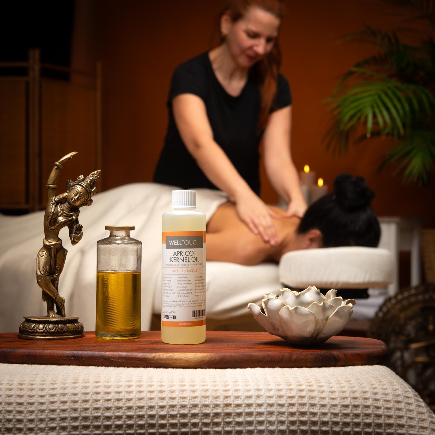 Welltouch Aprikosenkernöl-Flasche und Glasflasche mit Öl auf Holztablett. Frau erhält Massage. Dekoration.