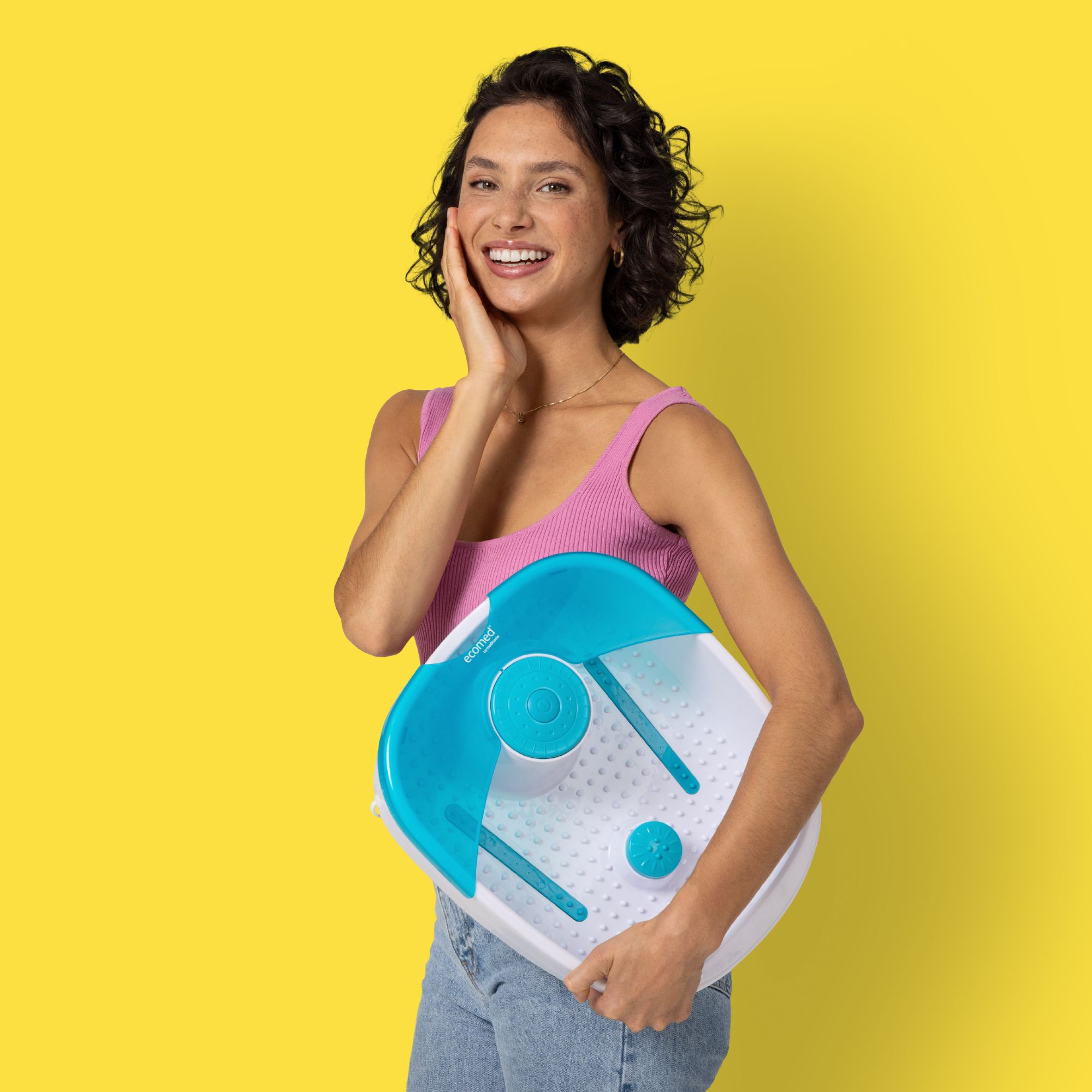 Frau hält Fußsprudelbad. Weiß-blaues Gerät. Frau lächelt, trägt rosa Top und Jeans. Gelber Hintergrund.
