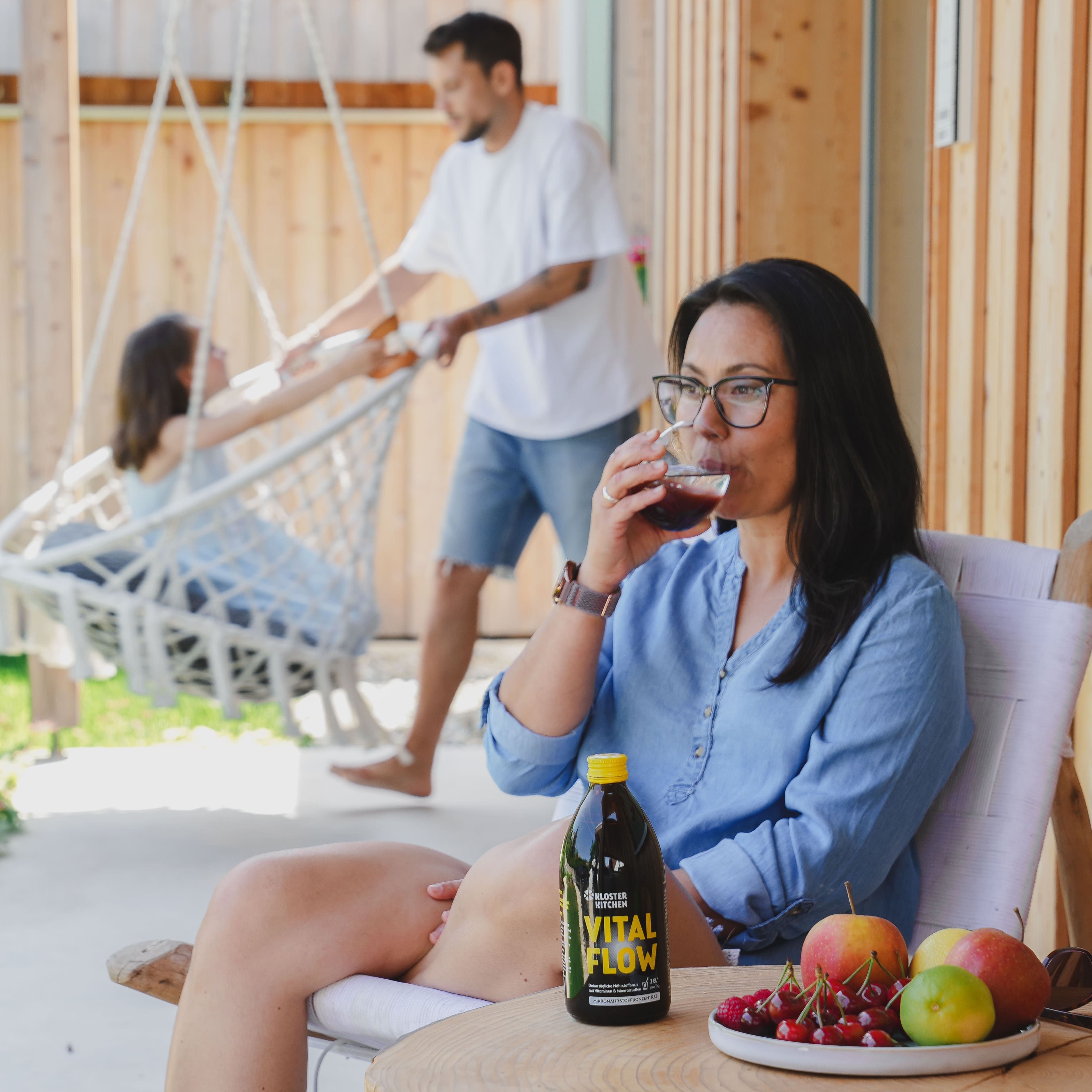 Frau trinkt aus einem Glas. Auf Tisch: Flasche VITAL FLOW, Obst. Im Hintergrund: Familie auf Schaukel.
