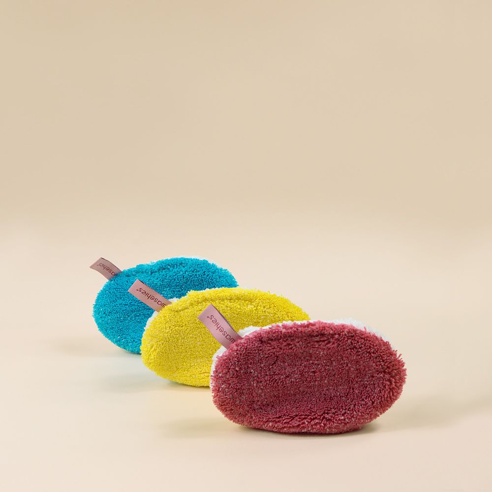 Drei farbige Waschpads mit Etikett, in Reihe angeordnet. Waschpads sind oval und haben eine Schlaufe.