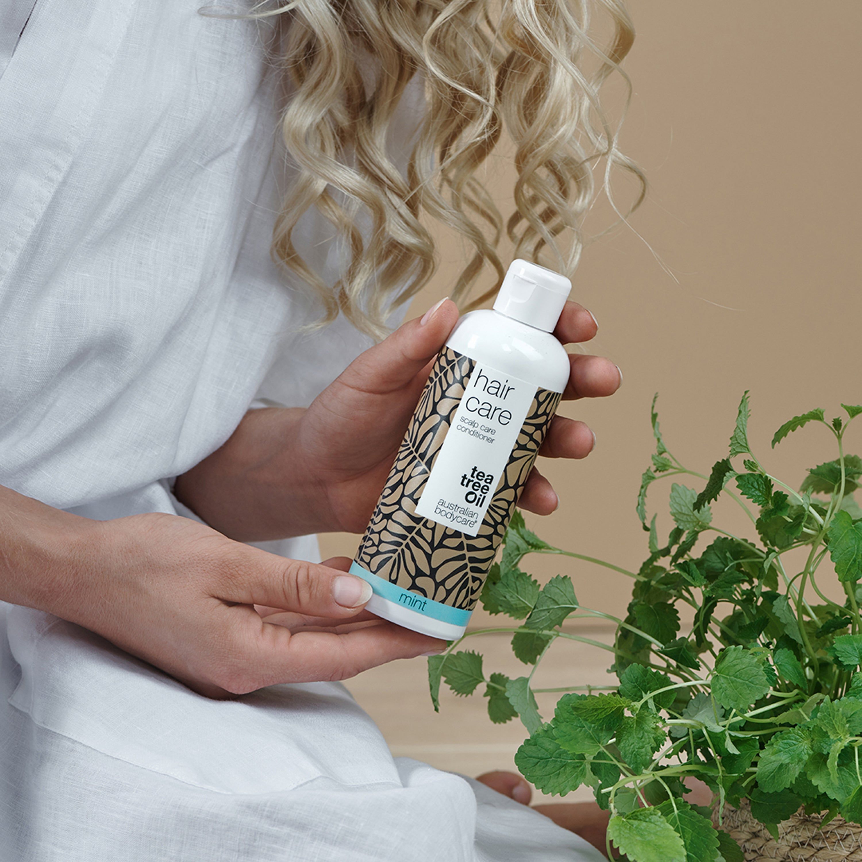 Frau hält eine Flasche vor einer Pflanze. Aufschrift "hair care", "tea tree oil" und "mint".