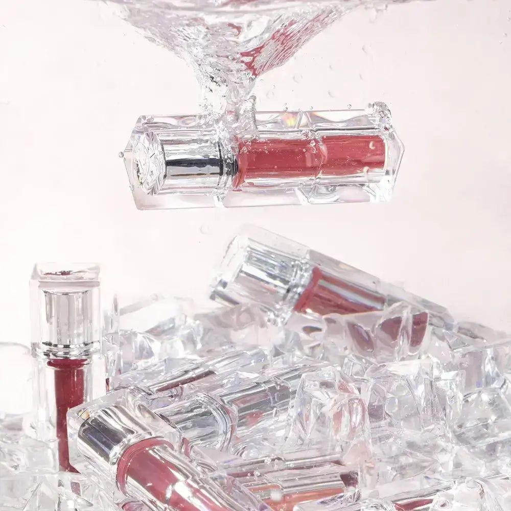 Mehrere durchsichtige Lippenstiftbehälter mit silbernen Kappen, einige in Eiswürfeln, einer fällt in Wasser.