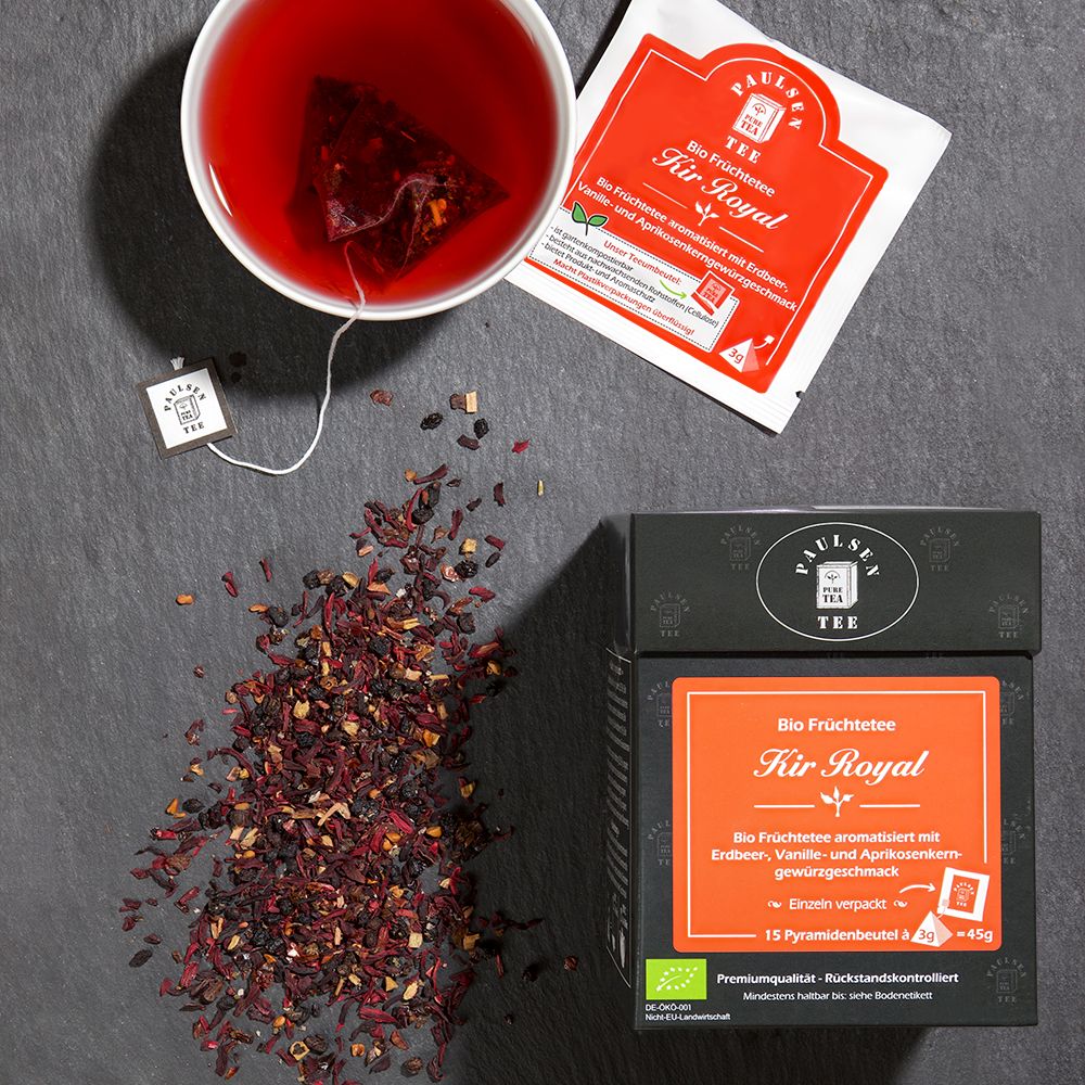 Tasse Tee und Teebeutel "Kir Royal" neben Tee-Box und losen Teeblättern. Box mit rotem Etikett, Pyramidenbeutel.