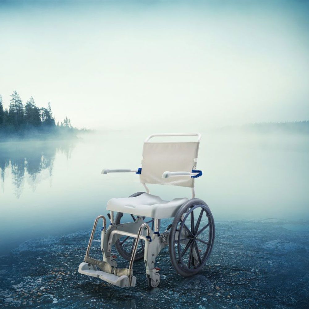 Dusch- und Toilettenrollstuhl am See. Weißer Rahmen, graue Räder, Sitz mit Öffnung. Armlehnen, Fußstützen. Blaue Akzente.