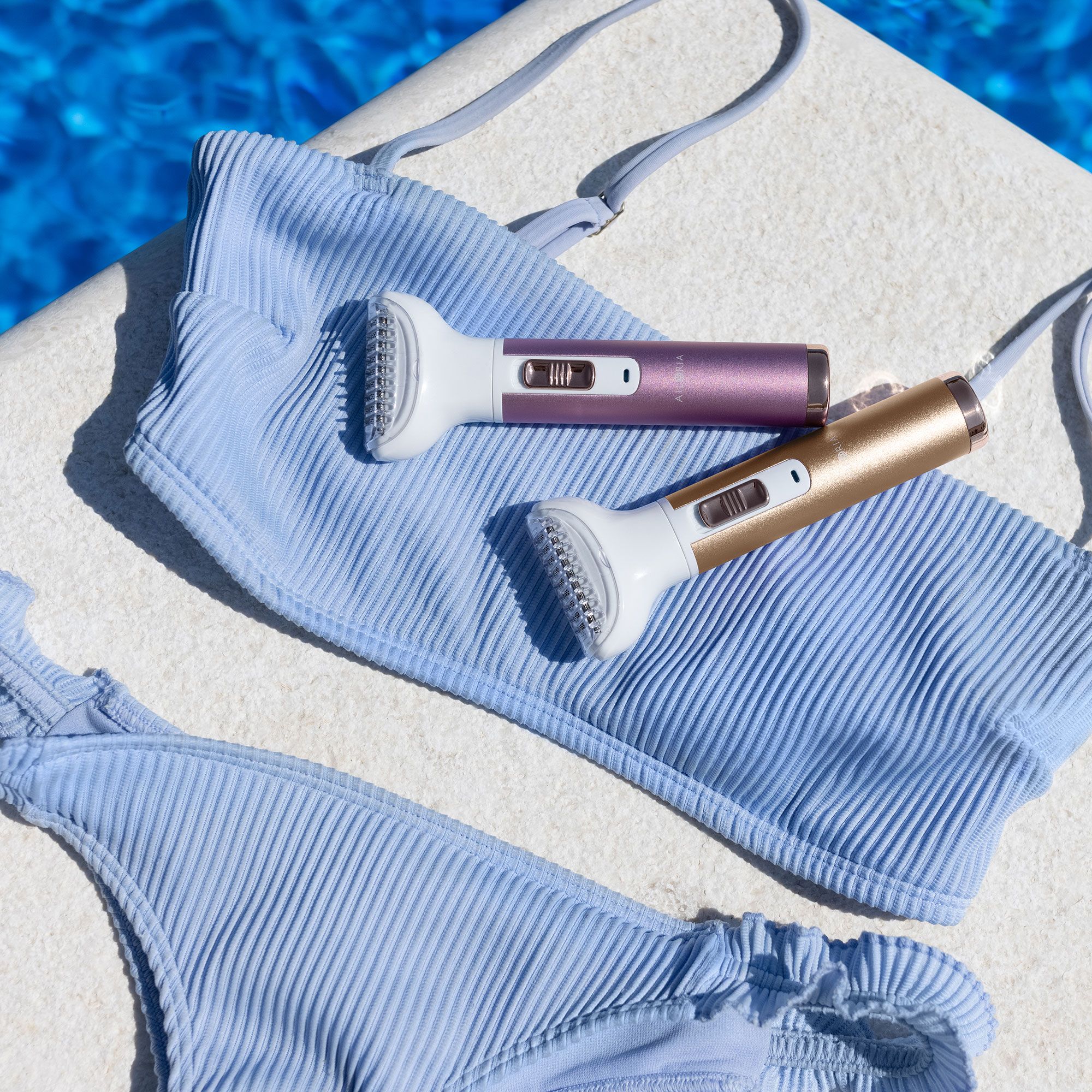 Zwei Rasierer mit weißen Köpfen und farbigen Griffen. Liegen auf einem blauen Bikini. Gold und Rosa.