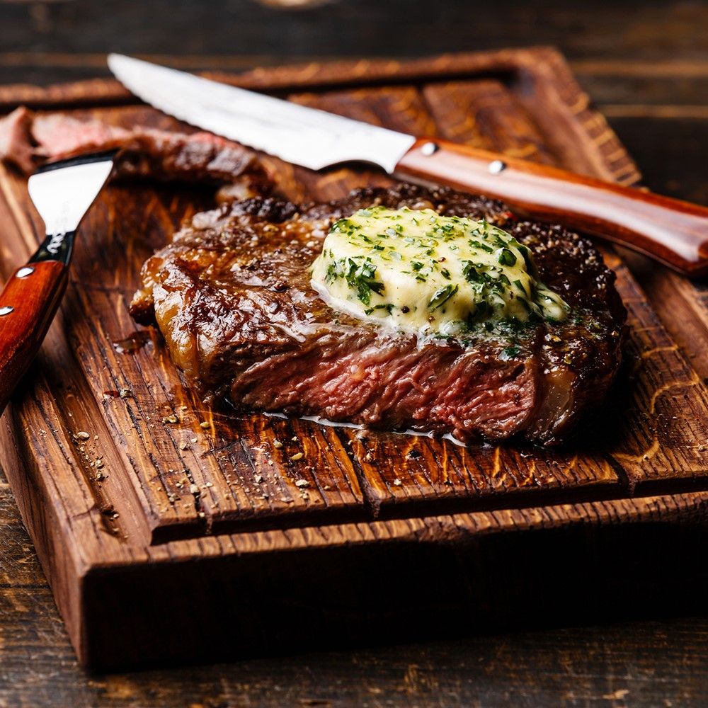 Gebratenes Steak auf Holzbrett. Kräuterbutter auf dem Steak. Messer und Gabel daneben. Holzbrett mit Rillen.