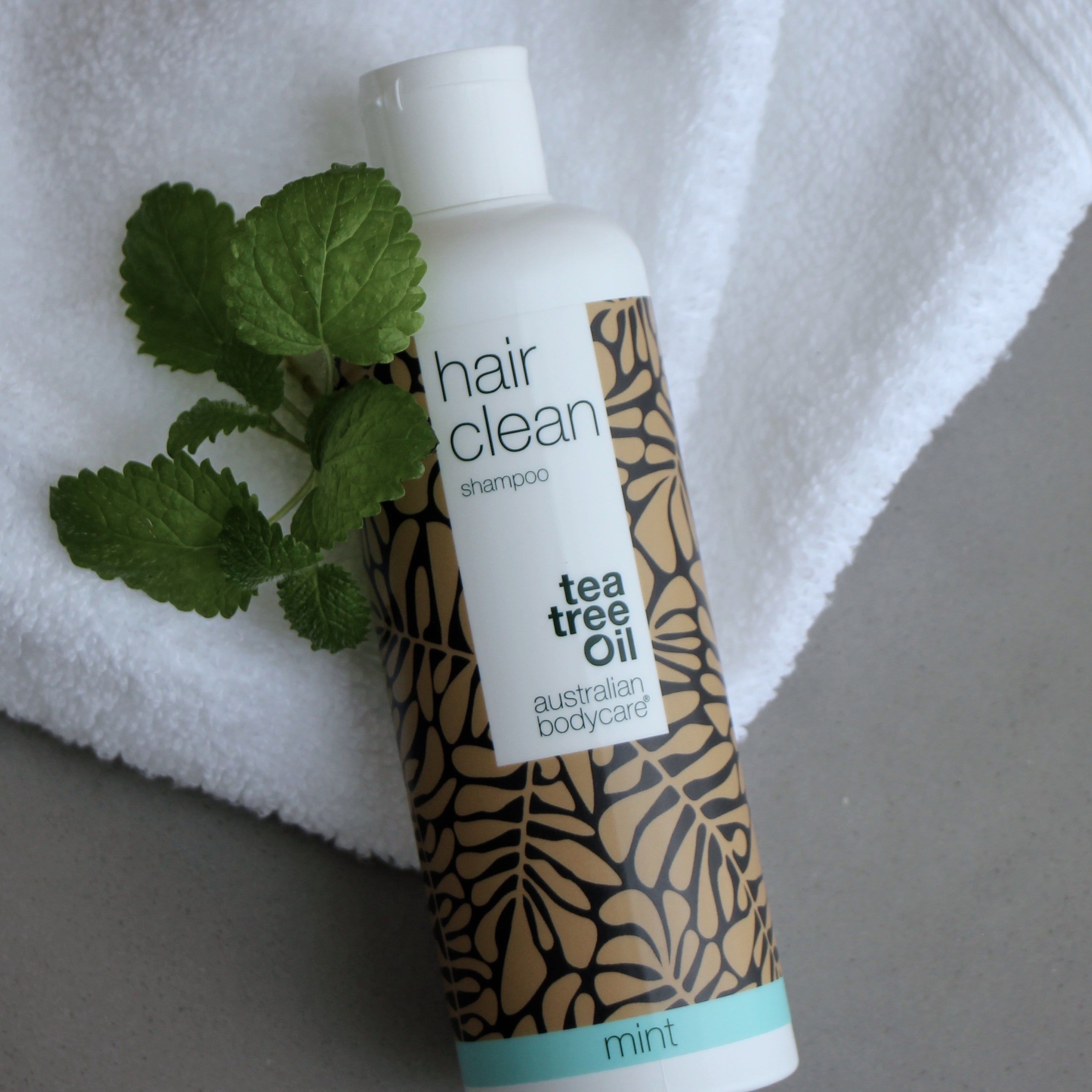 Shampoo-Flasche mit floralem Muster, Schriftzug 'hair clean shampoo', 'tea tree oil' und 'mint'. Auf Handtuch, daneben Minze.