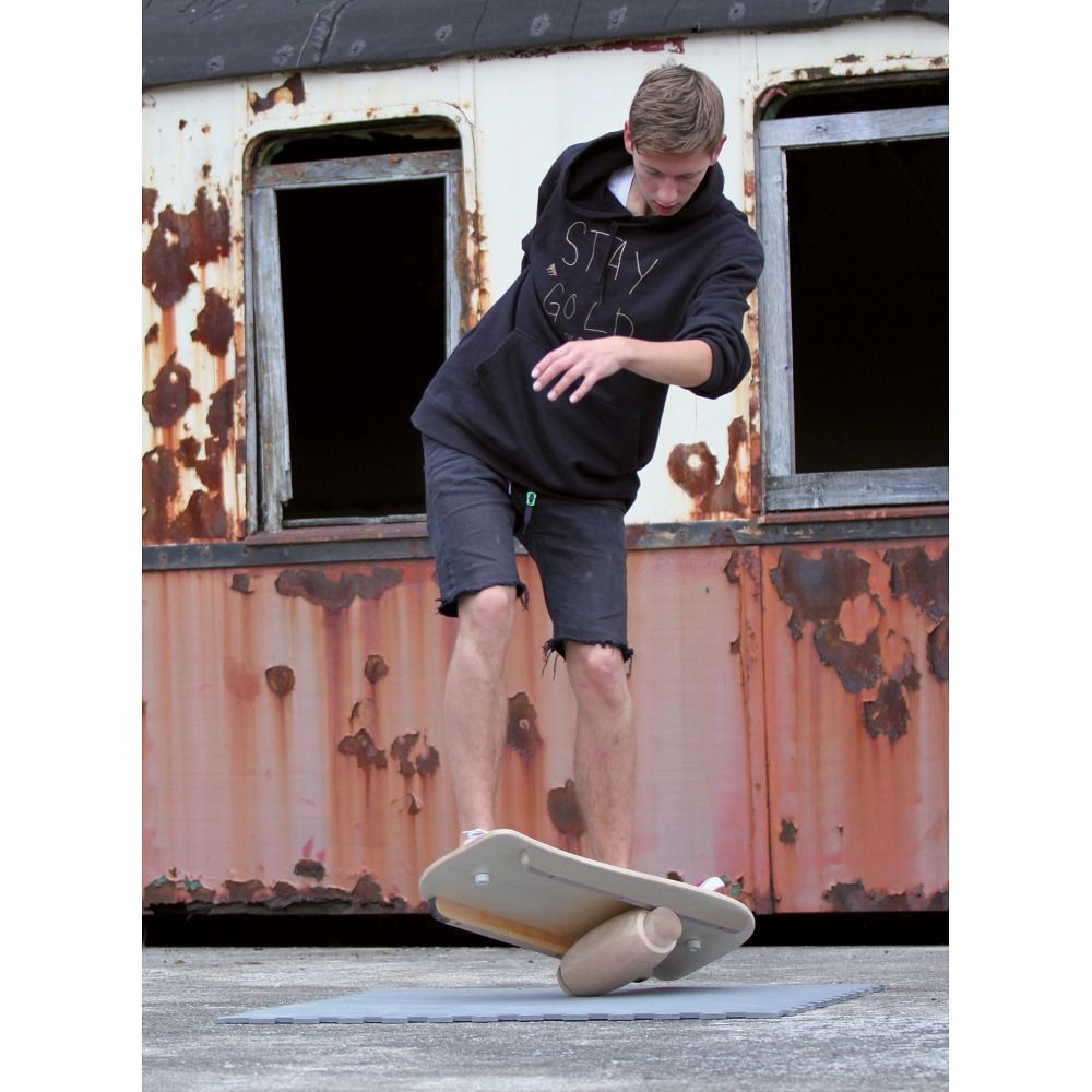 Mann balanciert auf einem Balance Board. Das Board liegt auf einer Rolle. Im Hintergrund ein rostiges Gebäude.