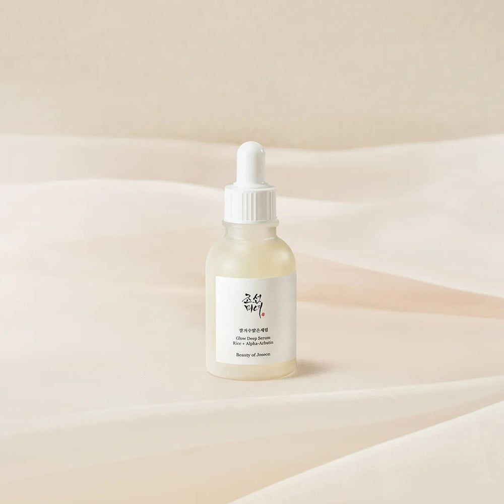 Serumflasche mit weißem Etikett. Beschriftung: Glow Deep Serum, Rice + Alpha-Arbutin. Marke: Beauty of Joseon. Auf hellem Stoff.