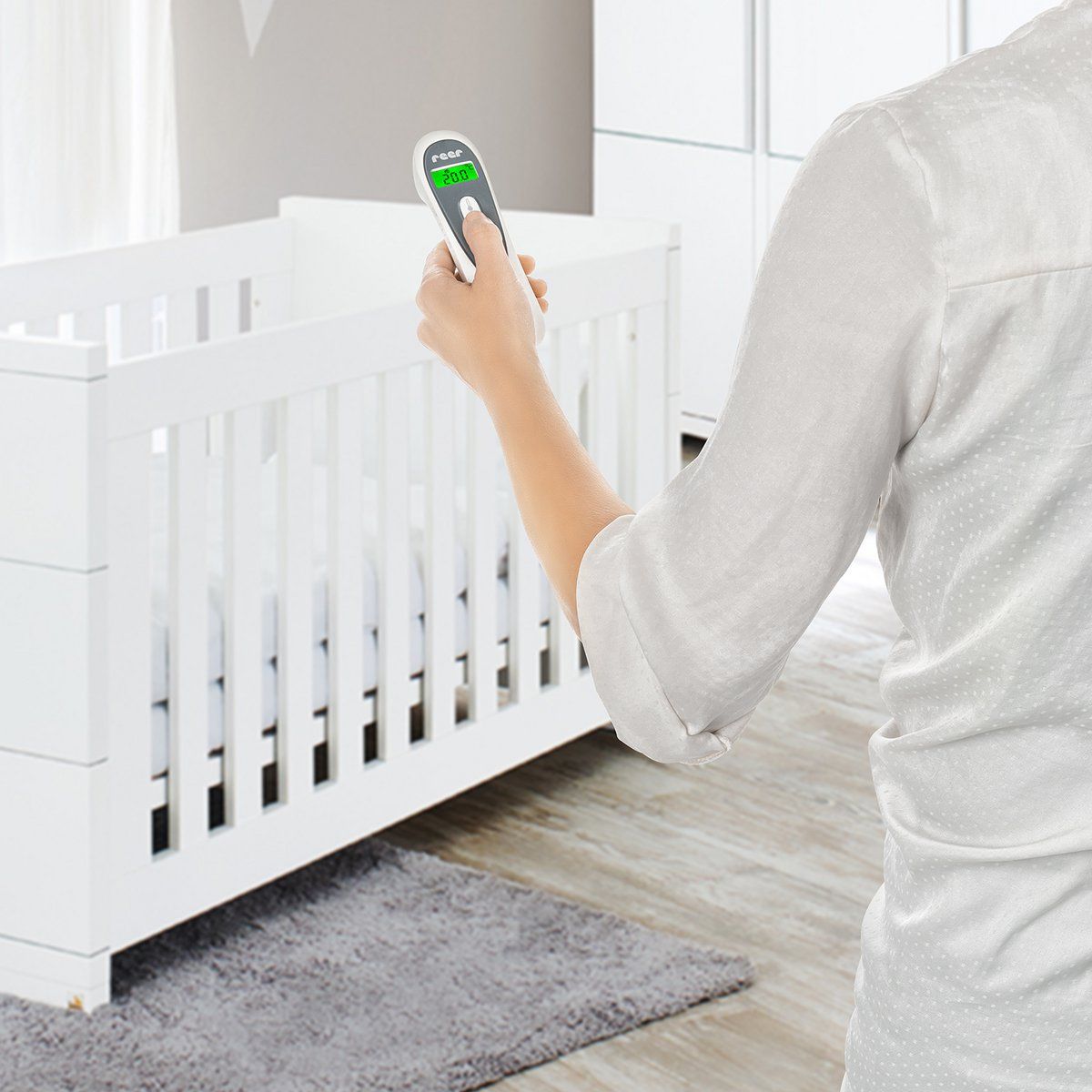 Person hält ein kontaktloses Infrarot-Thermometer. Marke: reer. Gerät wird auf ein Babybett gerichtet.