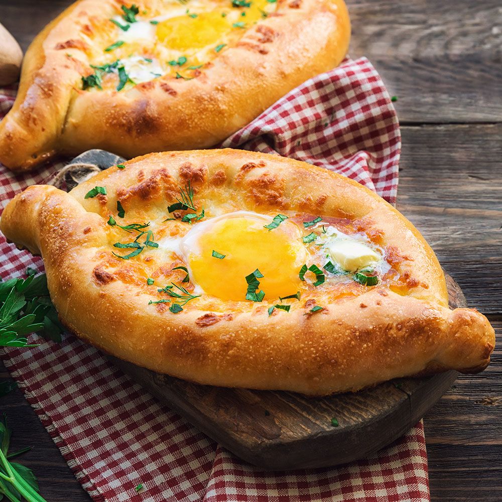 Zwei Khachapuri-Brote mit Eiern. Goldbrauner Teig, gelbes Ei. Auf Holzbrett und rot-weiß kariertem Tuch.