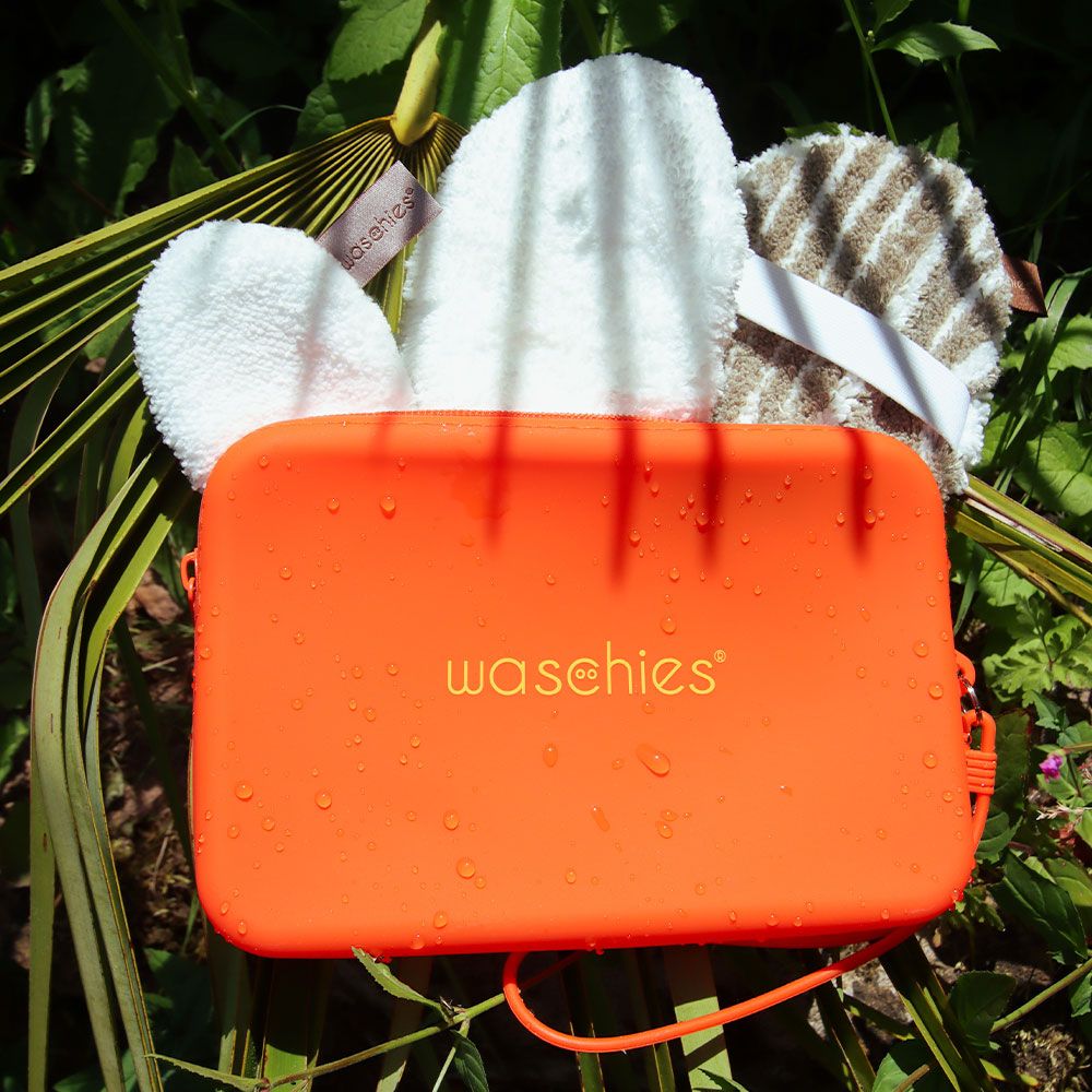 Orangefarbene Tasche mit Reißverschluss und Griff, liegt auf Blättern. Enthält drei runde Waschpads. Auf der Tasche steht "waschies".