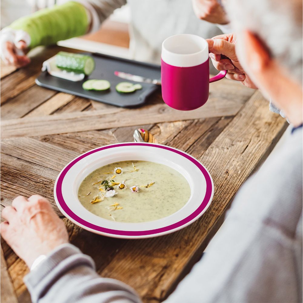 Teller mit Suppe, weiß mit pinkfarbenem Rand. Person hält Tasse. Auf Holztisch. Gurken und Messer auf Tablett.