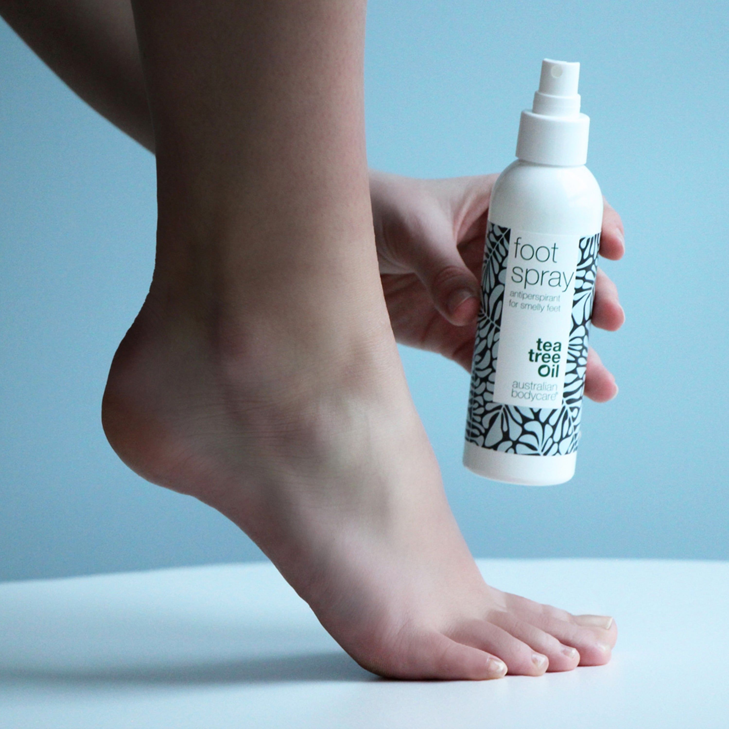 Fußspray-Flasche mit weißem Sprühkopf. Aufschrift: "foot spray" und "tea tree oil". Hält eine Person die Flasche neben einem Fuß.