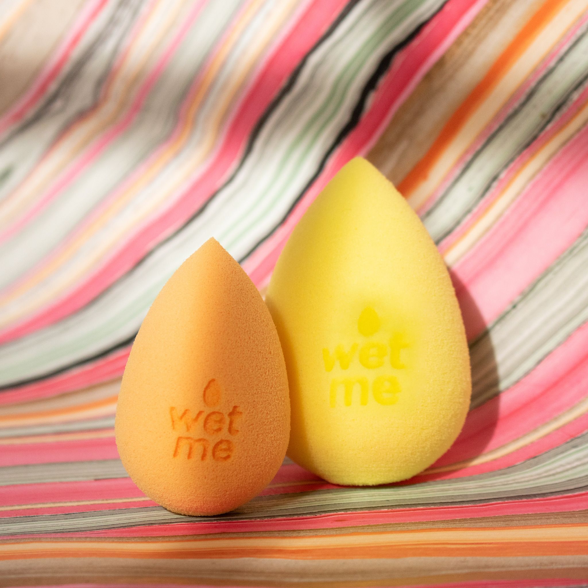 Zwei Beautyblender Make-up-Schwämme, orange und gelb, auf gestreiftem Hintergrund.