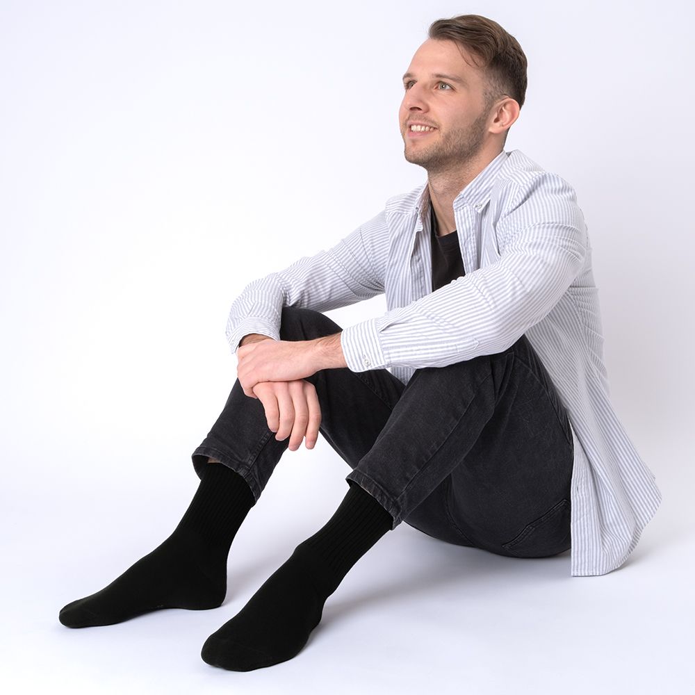 Mann sitzt mit schwarzen Socken ohne Gummibund. Gestreiftes Hemd, dunkle Hose.