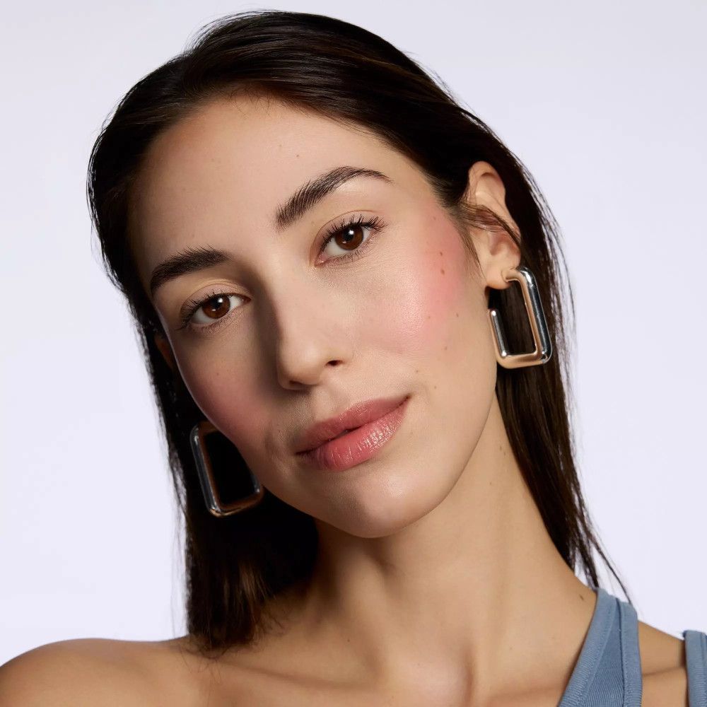 Porträt einer Frau mit rosa Rouge auf den Wangen. Sie trägt Ohrringe und hat einen neutralen Gesichtsausdruck.