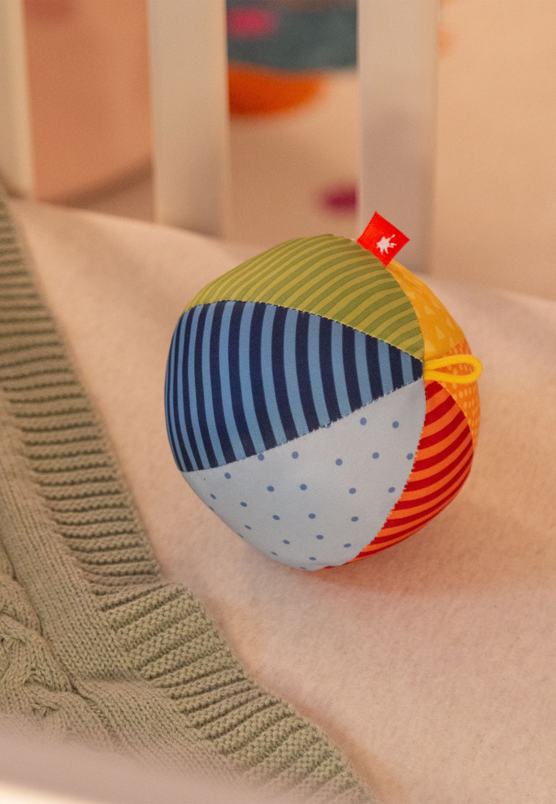 Bunter Stoffball mit gelbem Aufhänger. Rotes Etikett mit Logo. Teilweise im Hintergrund.
