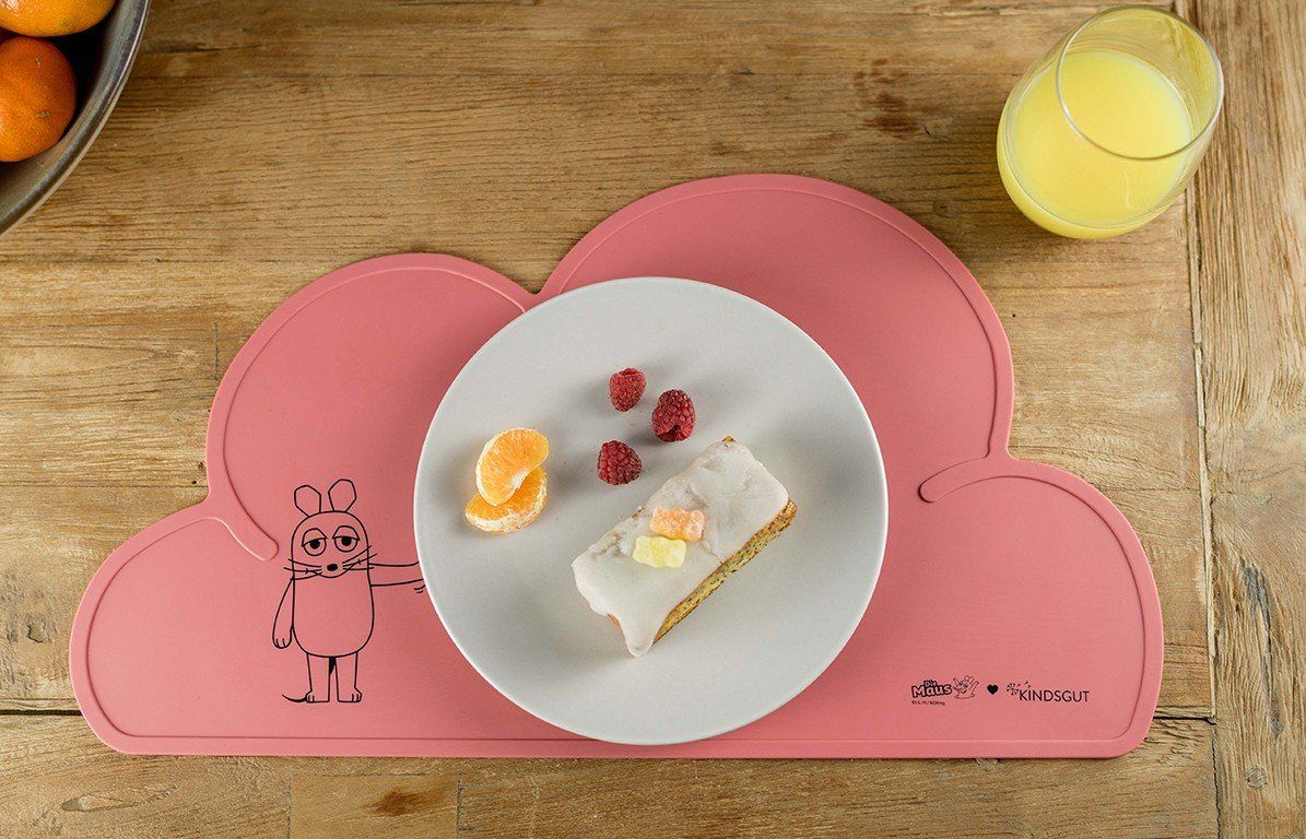 Rosa Platzdeckchen in Wolkenform mit Essen. Teller mit Kuchen, Obst und Glas Orangensaft.
