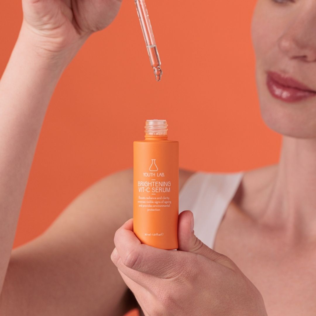 Frau hält Flasche und Pipette. Flasche orange, Aufschrift: YOUTH LAB Brightening Vit-C Serum. Hintergrund orange.