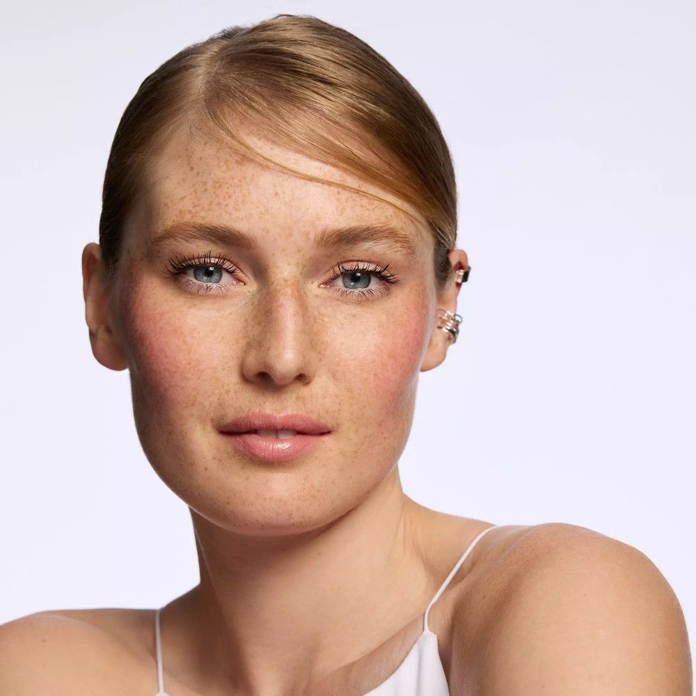 Porträt einer Frau mit rosa Rouge auf den Wangen. Sie trägt ein weißes Oberteil. Ihr Haar ist blond und hat Sommersprossen.