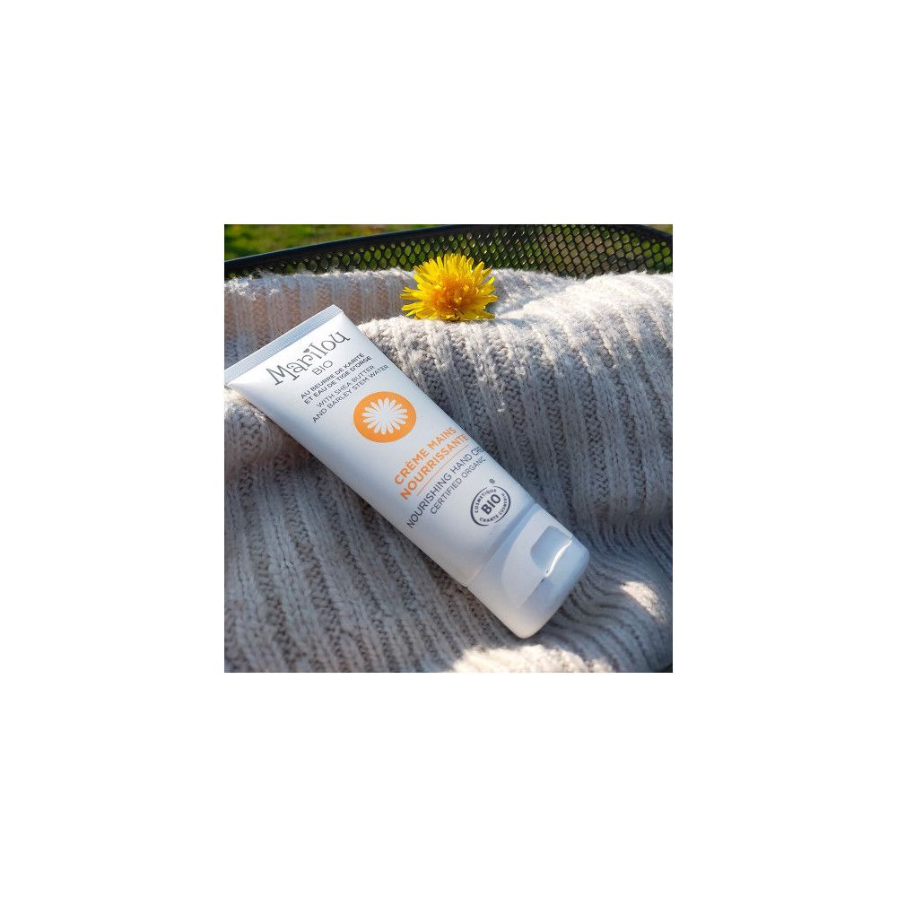 Handcreme-Tube mit Marilou Bio Logo. Liegt auf einem grauen Pullover, mit Blume.