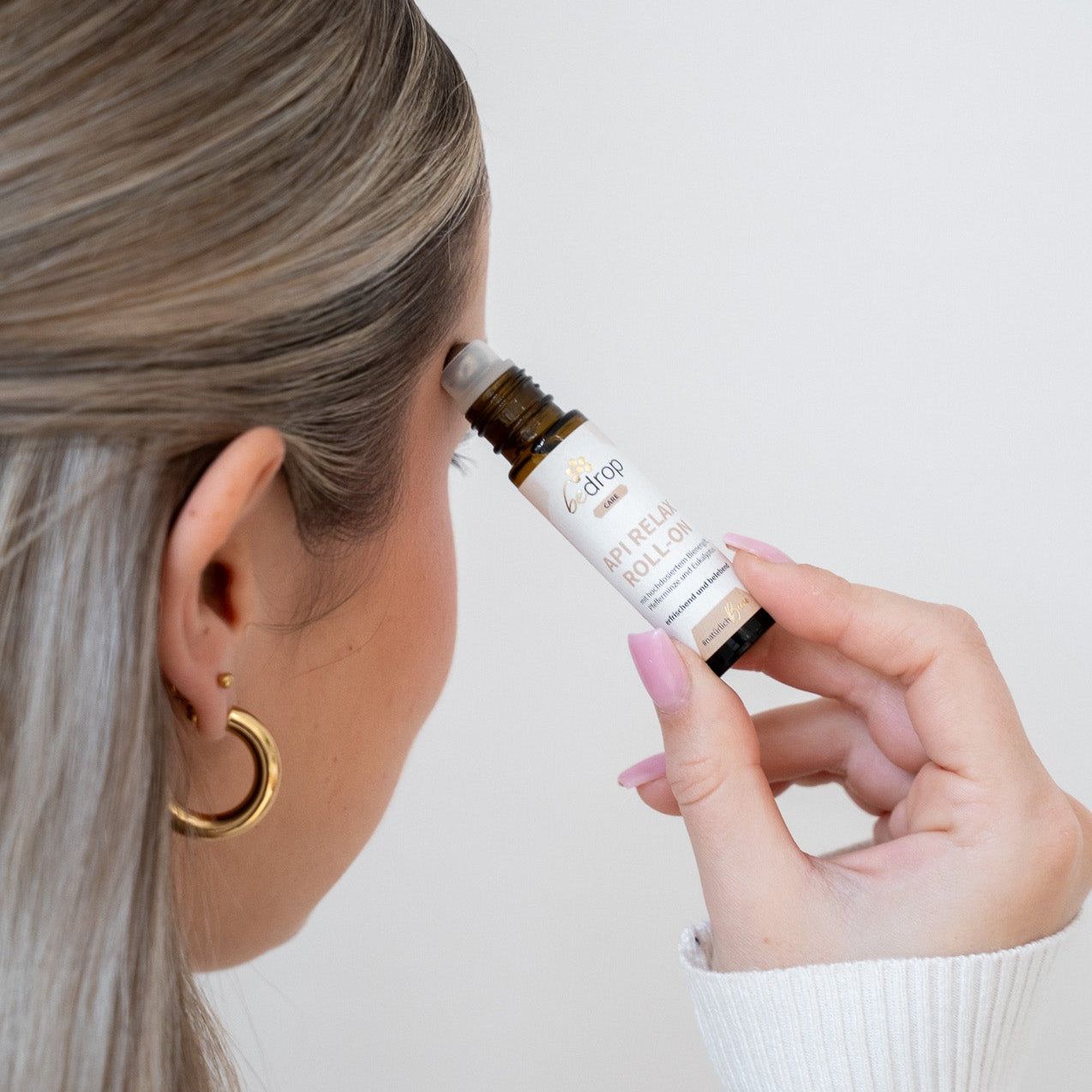 Person hält braune Glasflasche mit schwarzem Deckel. Aufschrift: bedrop API RELAX ROLL-ON. Anwendung am Kopf.