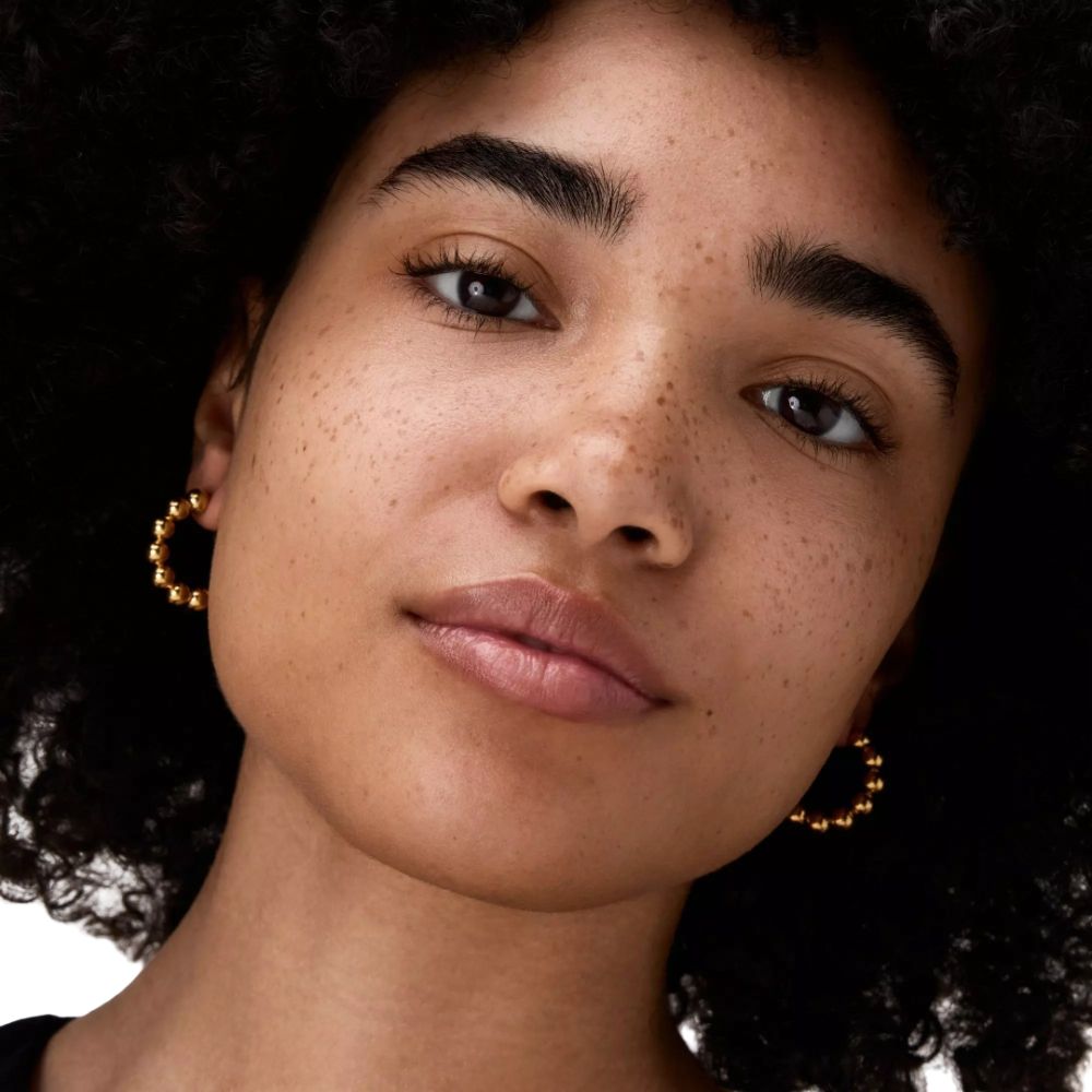 Porträt einer Frau mit Sommersprossen. Sie trägt goldene Ohrringe.