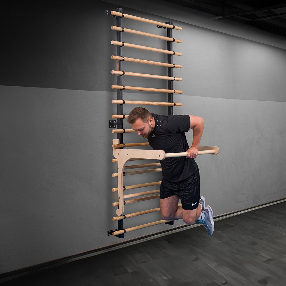 Mann trainiert an Klimmzugbügel Multi. Sprossenwand im Hintergrund. Schwarze Kleidung.