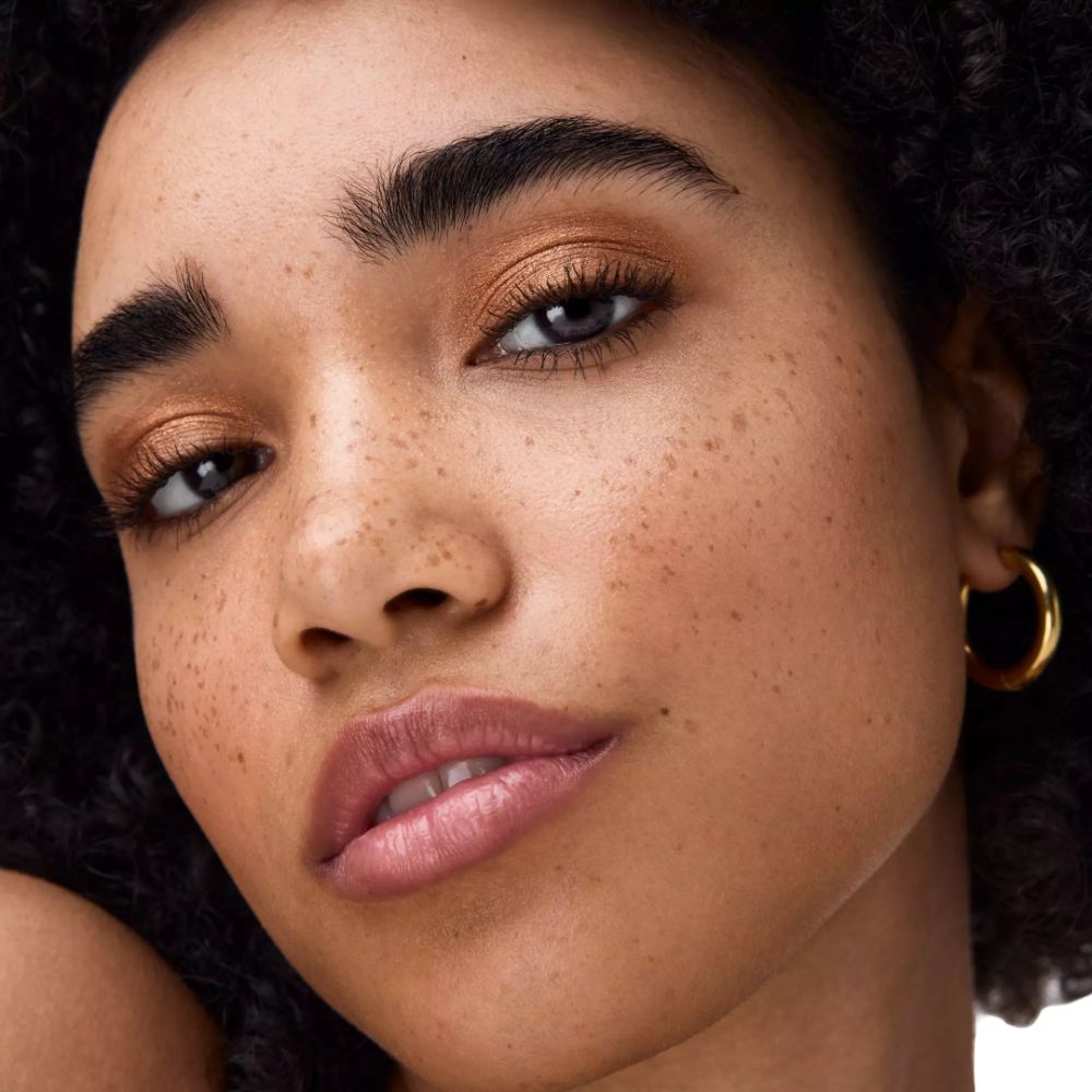 Nahaufnahme eines Gesichts mit goldfarbenem Lidschatten. Dunkler Hautton, Sommersprossen, goldene Creolen.