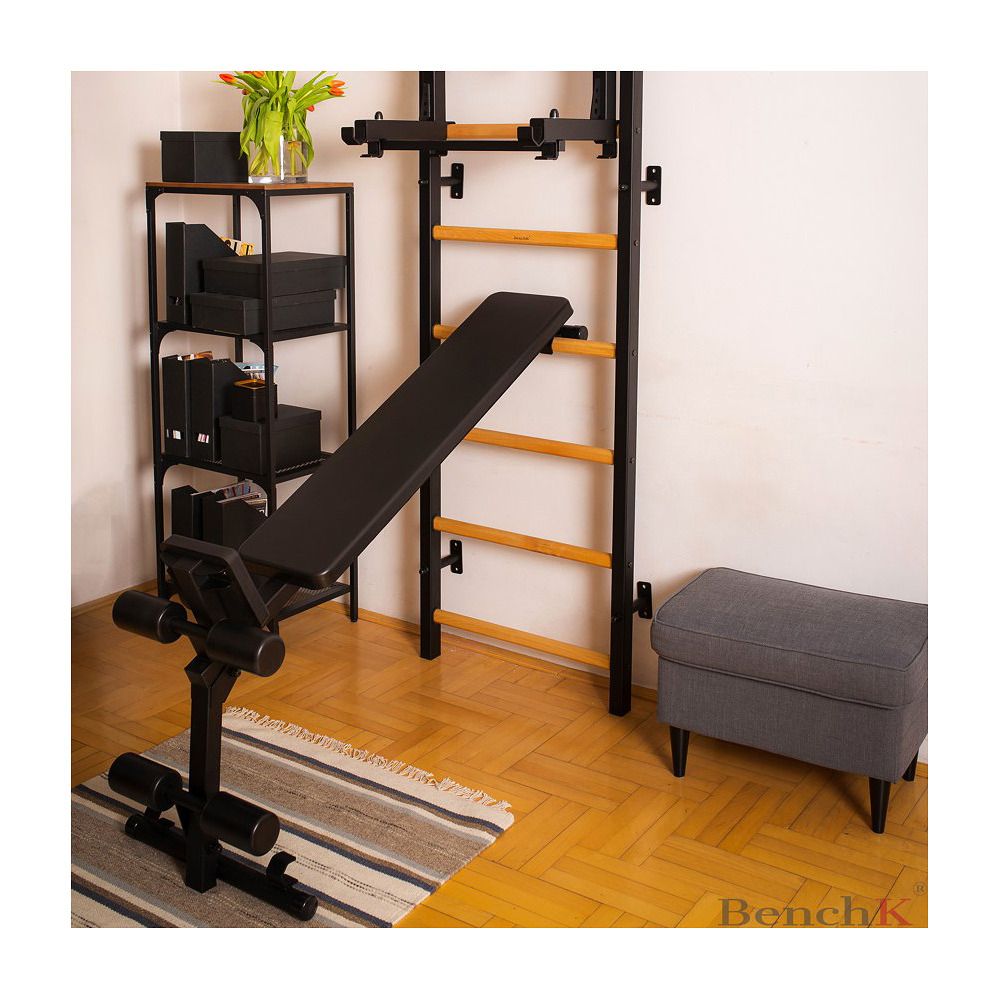 Schwarze Trainingsbank mit Sprossenwand und Zubehör. Metallrahmen. Grauer Hocker und Dekoration im Hintergrund. BenchK Logo.