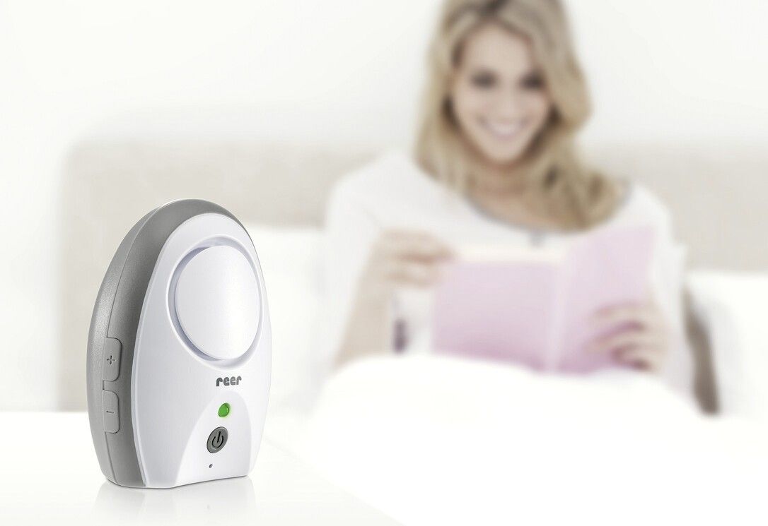Babyphone Neo, weiß-grau. Ein-/Ausschalter und grüne LED. Liegt auf einem Bett, im Hintergrund eine Frau. Marke: reer.