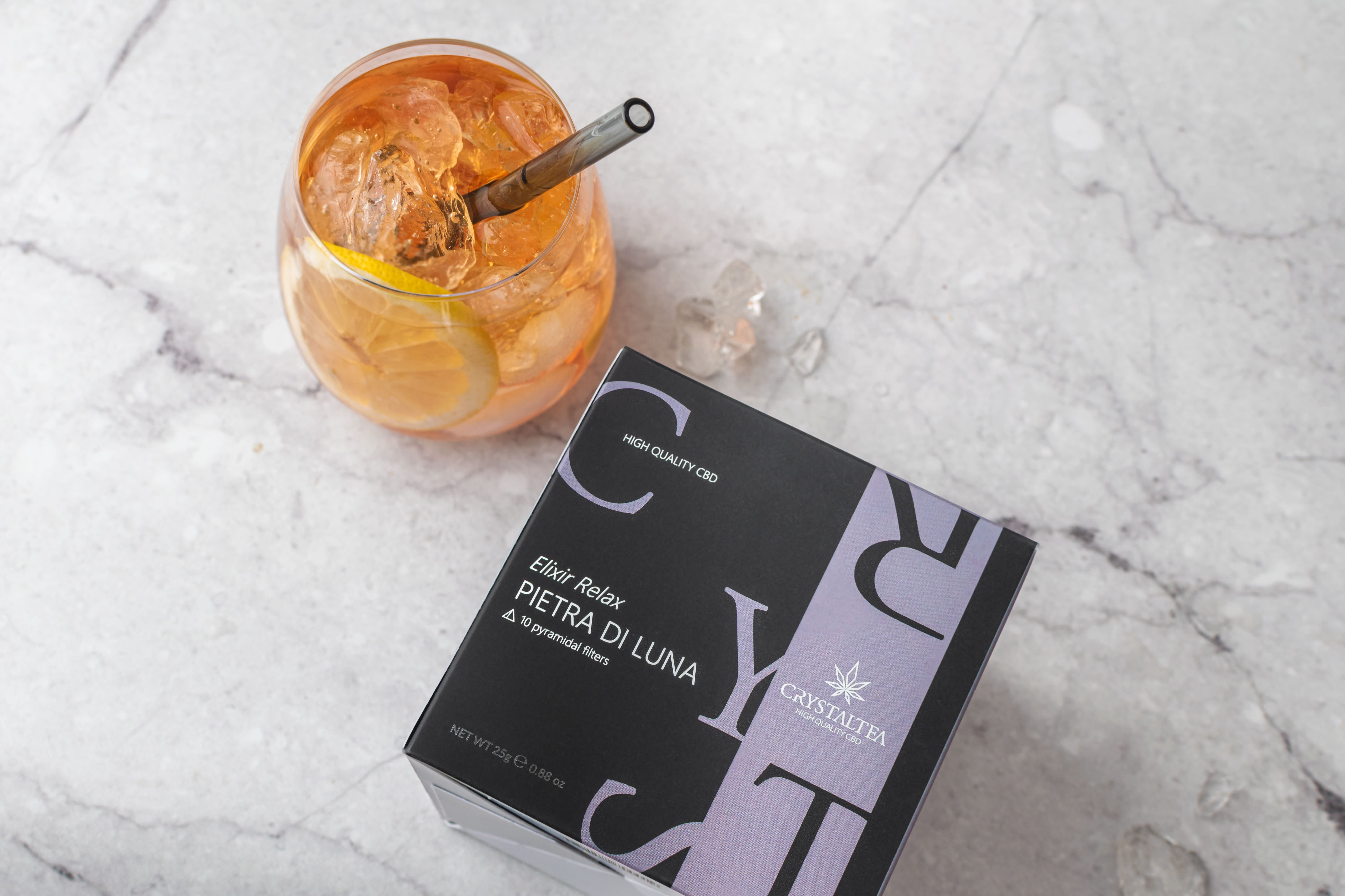 Glas mit Getränk, Eiswürfeln und Strohhalm. Daneben Schachtel mit Produktnamen und Logo.