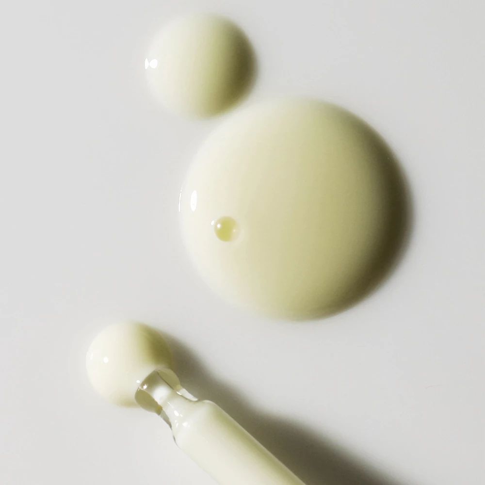 Drei Tropfen Serum, darunter ein Tropfen mit einer Pipette. Flüssigkeit ist hellgelb. Pipette mit weißem Griff.