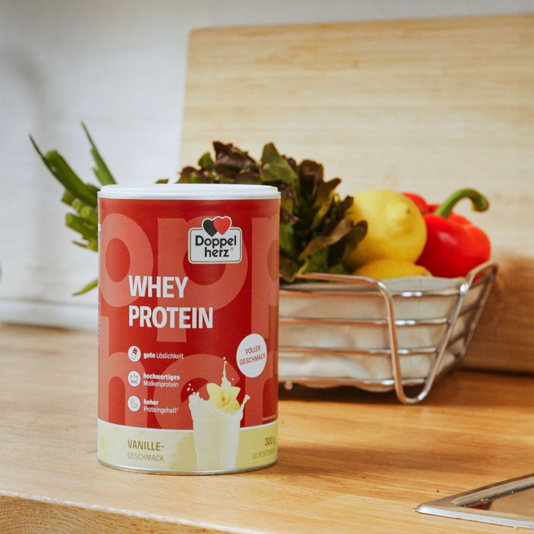 Dose Doppelherz WHEY PROTEIN Vanille. Rote Dose mit Logo und Produktinformationen. Daneben Gemüse und Obst.