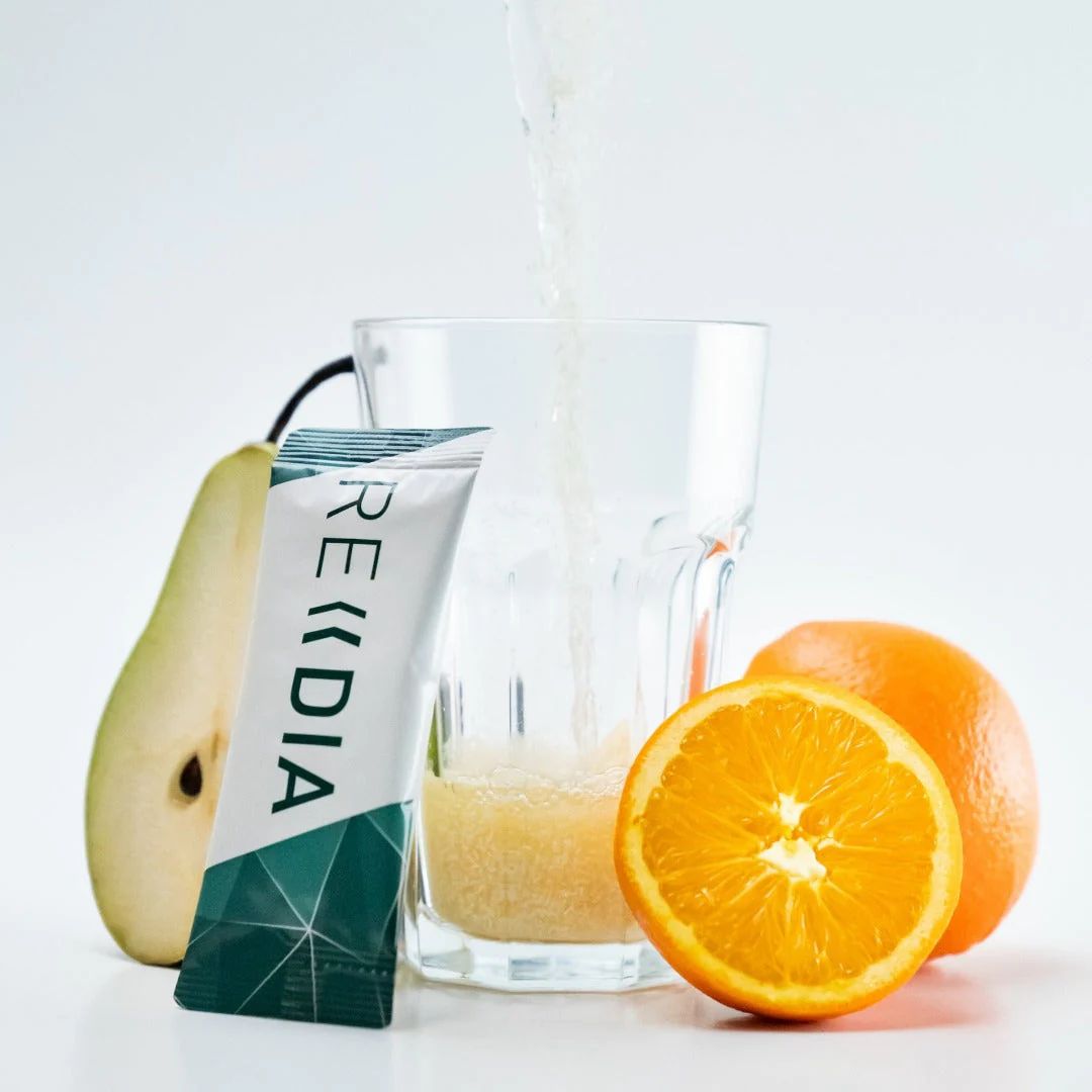 Beutel REDIA neben Glas mit Flüssigkeit, Birne und Orange. Flüssigkeit wird in Glas gegossen. Beutel mit grünem geometrischen Muster.