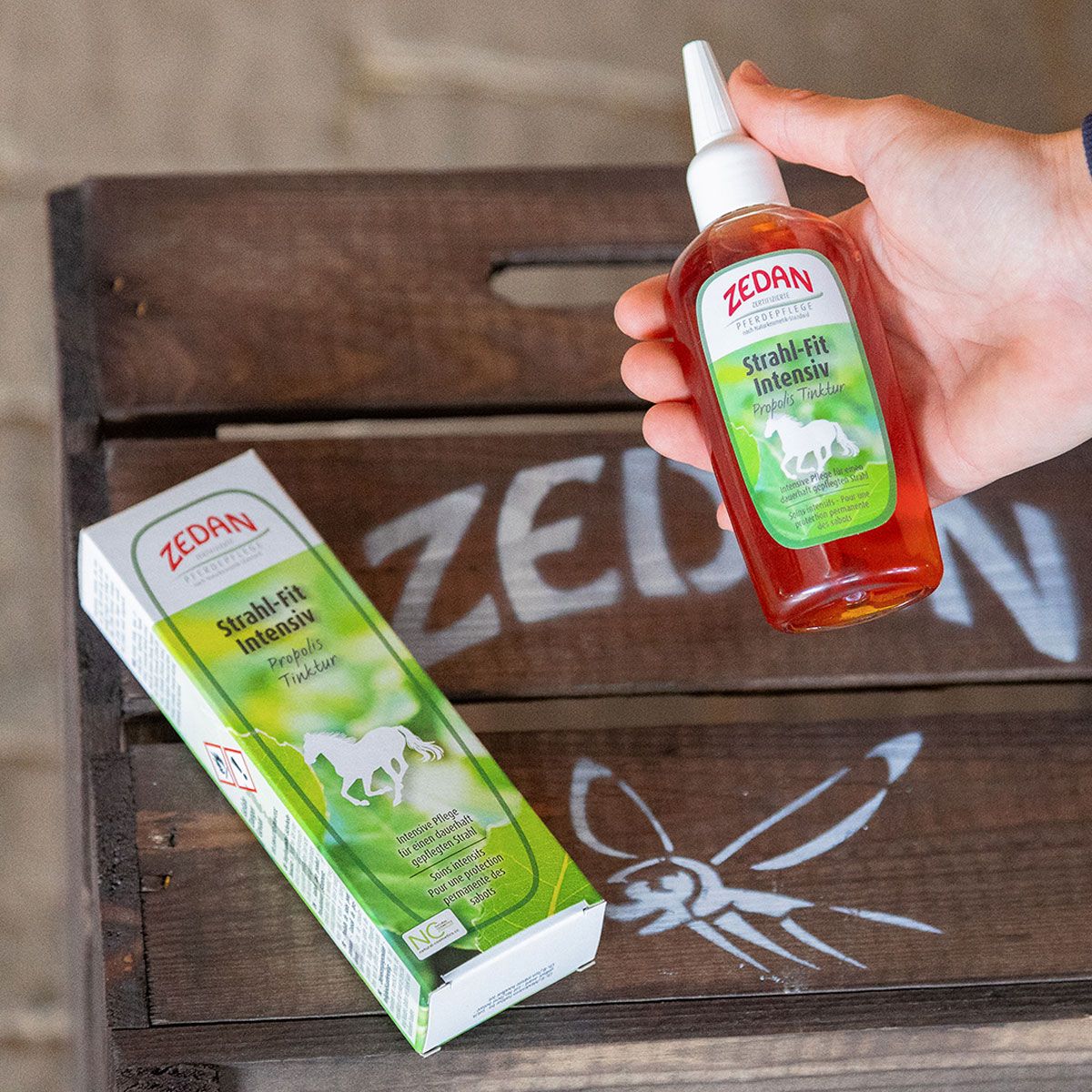 Hand hält Zedan Strahl-Fit Intensiv Flasche neben der Verpackung auf Holzkiste.