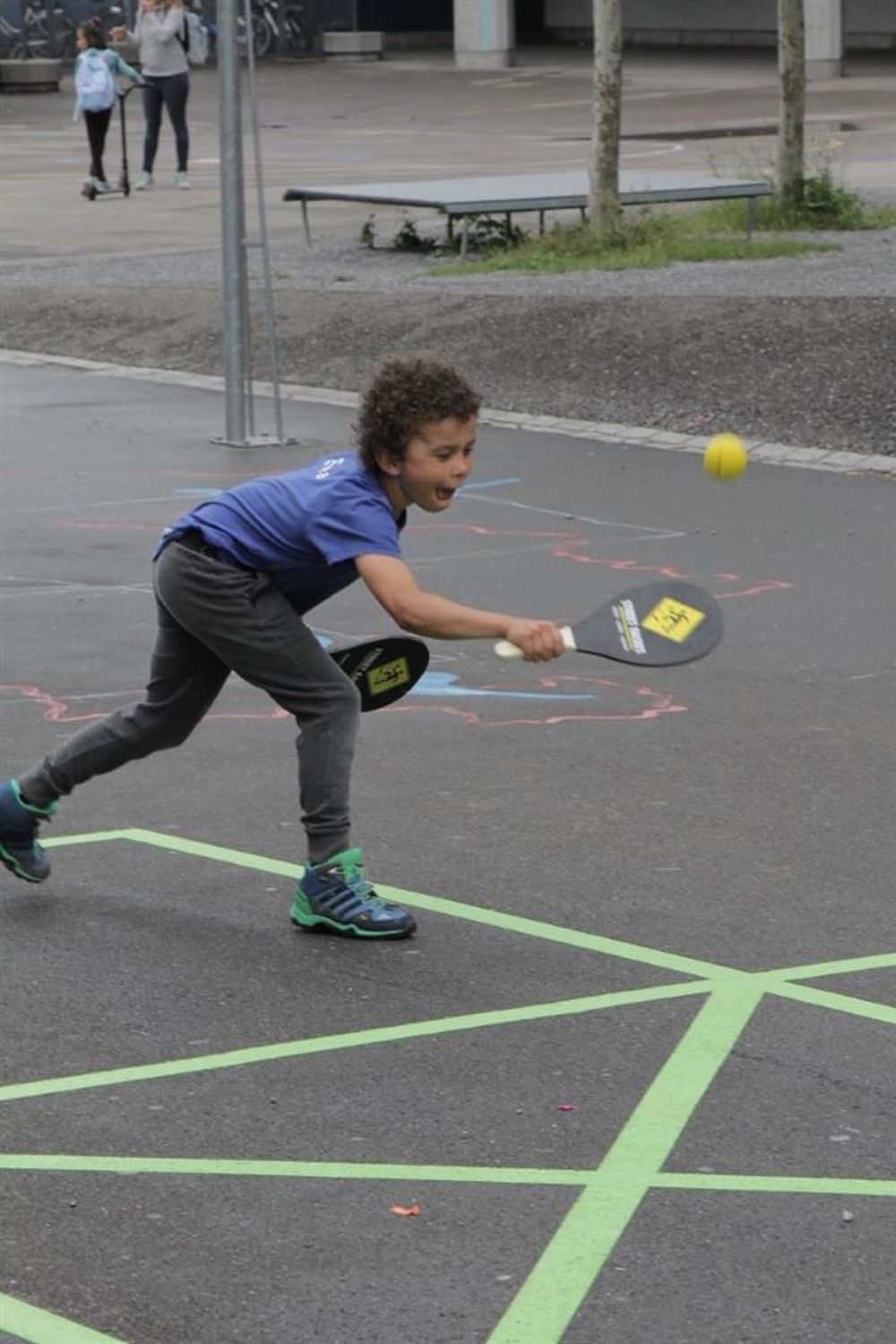 Kind spielt Street Racket. Schwarzer Schläger mit gelbem Logo. Kind trägt blaues T-Shirt und graue Hose. Spielt auf Asphalt.
