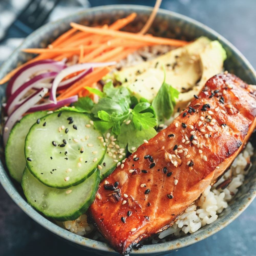 Schüssel mit Lachs, Reis, Avocado, Gurke, Karotten und Zwiebeln. Sesamkörner auf dem Lachs.