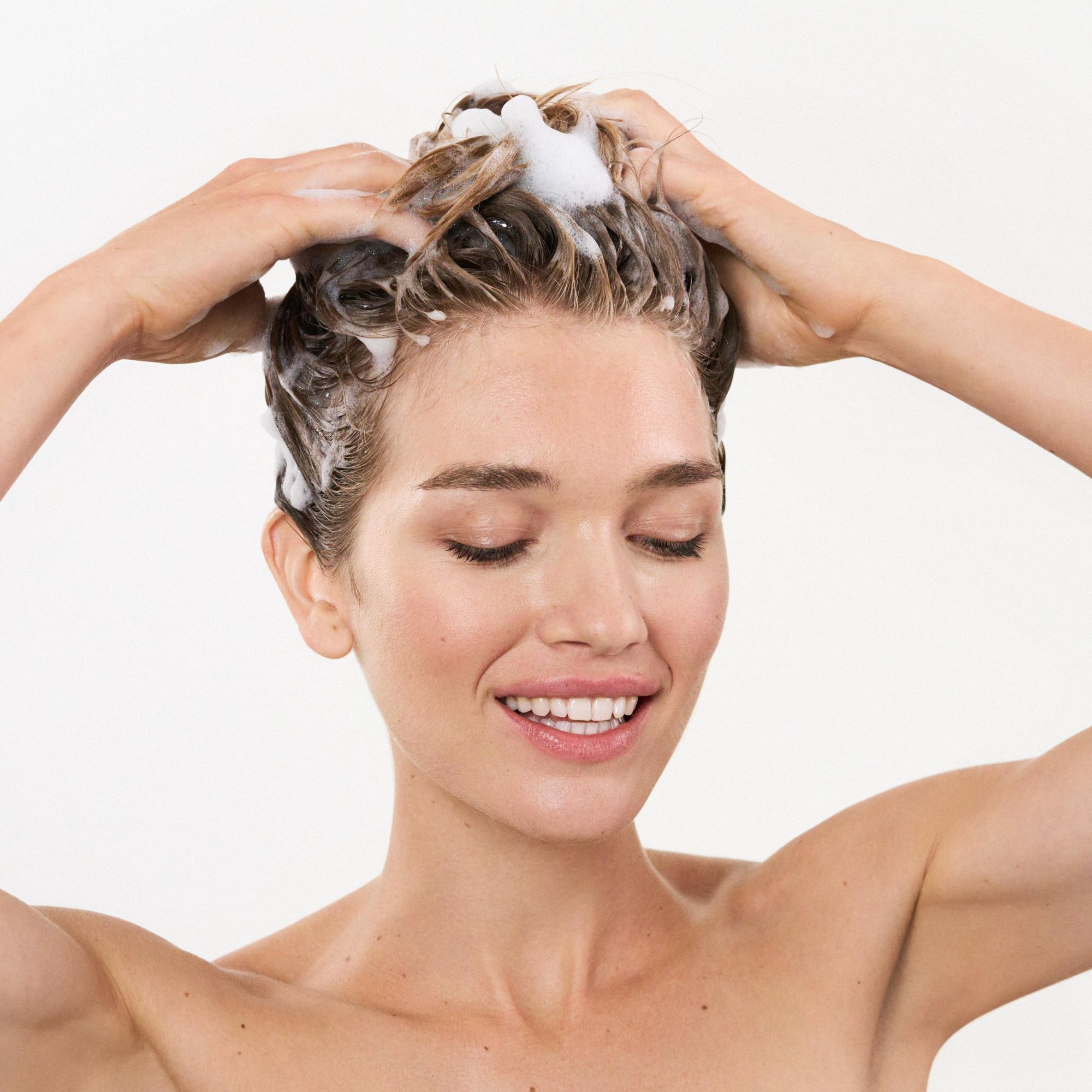 Frau wäscht sich die Haare mit Shampoo. Schaum auf dem Haar. Augen geschlossen, lächelnd.