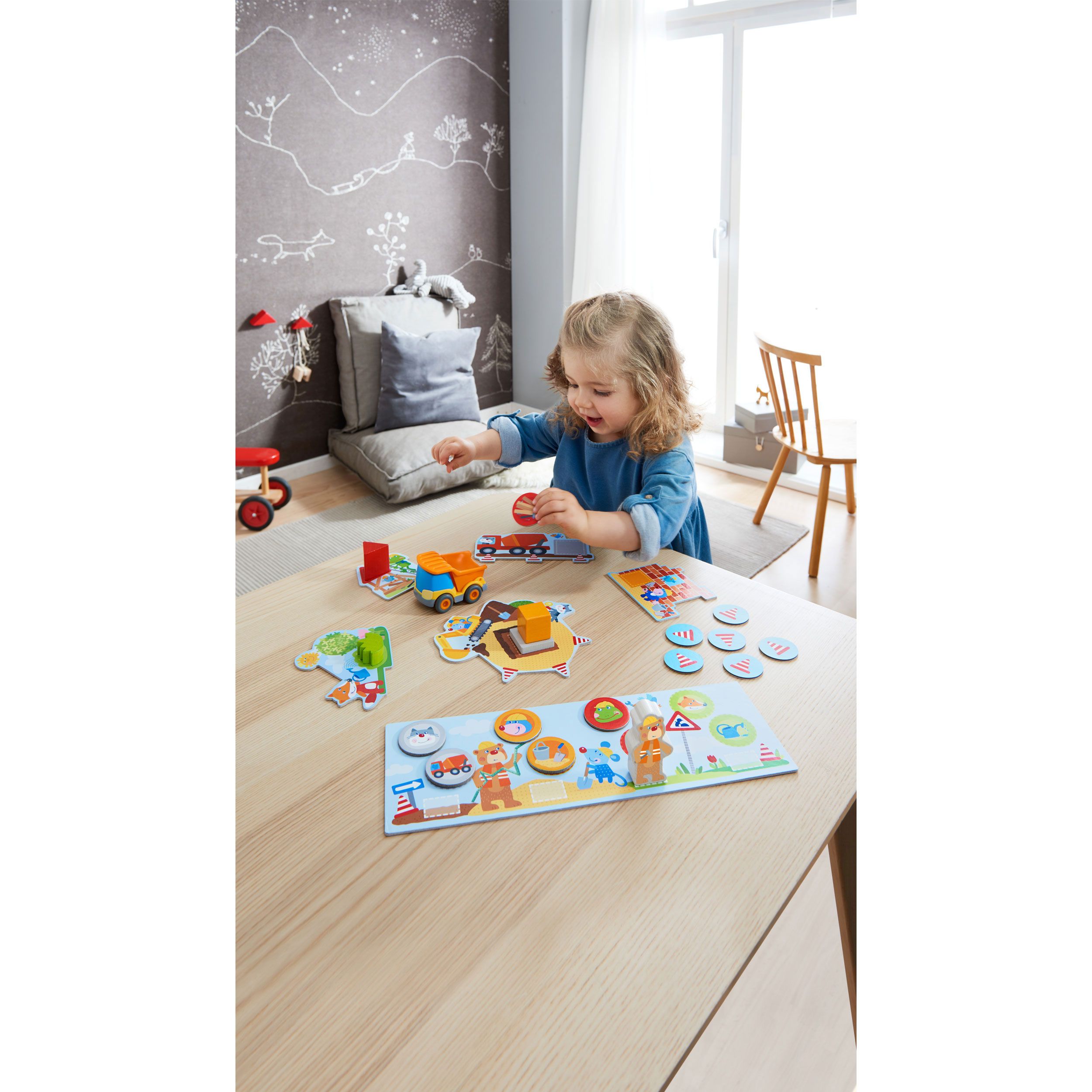 Kind spielt mit Baustellenspiel. Holzspielzeug, Puzzleteile, Spielfiguren und ein LKW auf einem Tisch. Hintergrund: Zimmer.