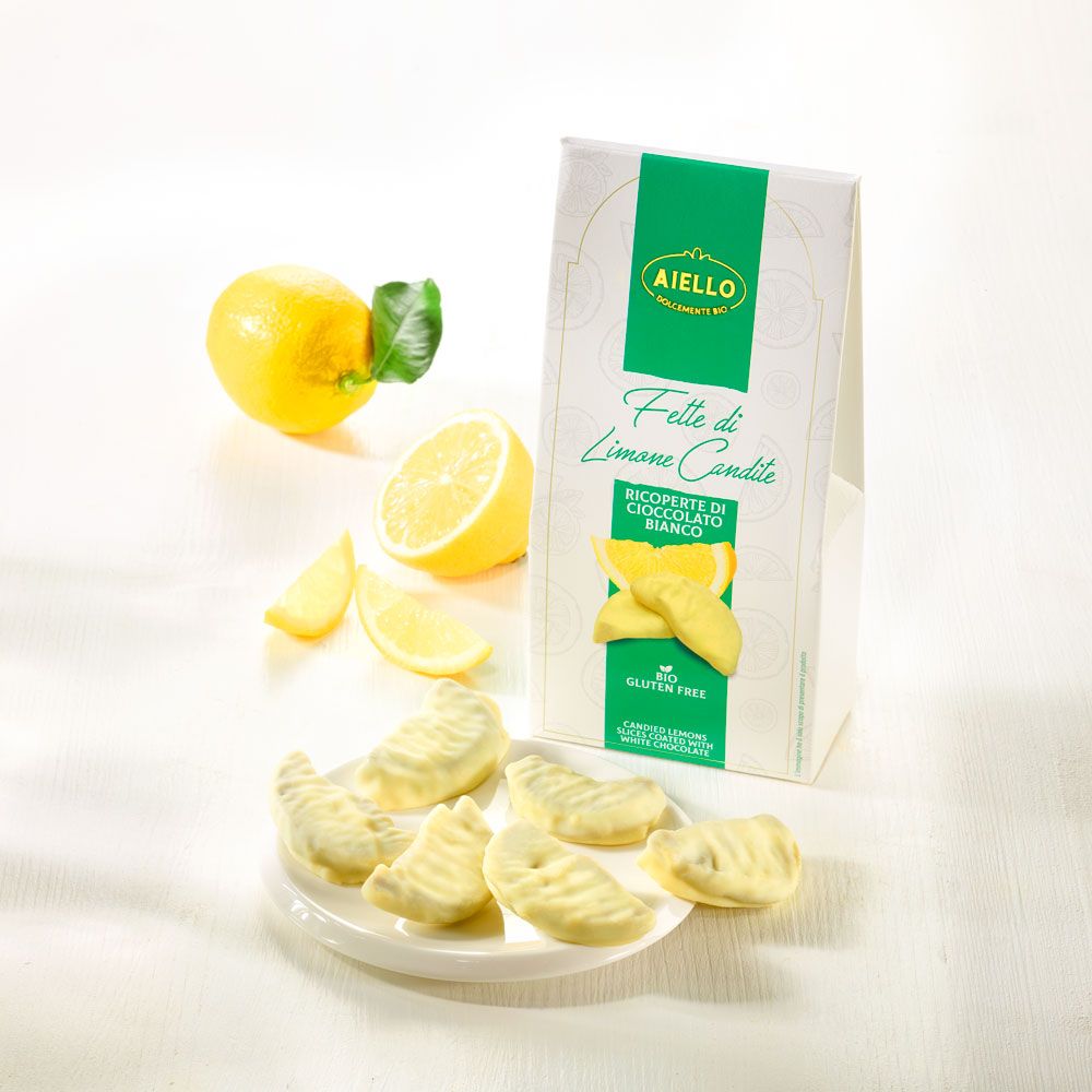 Verpackung und Teller mit kandierten Zitronenscheiben in weißer Schokolade. Zitrone und Scheiben daneben. Text: "AIello", "Fette di Limone Candite".