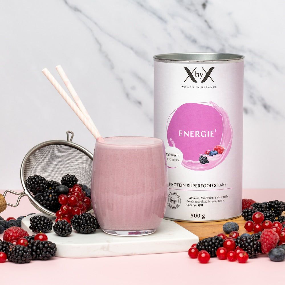 Glas mit Shake, Dose und Beeren. Dose mit „Energie“ und „Protein Superfood Shake“. Beeren und Sieb auf Unterlage.