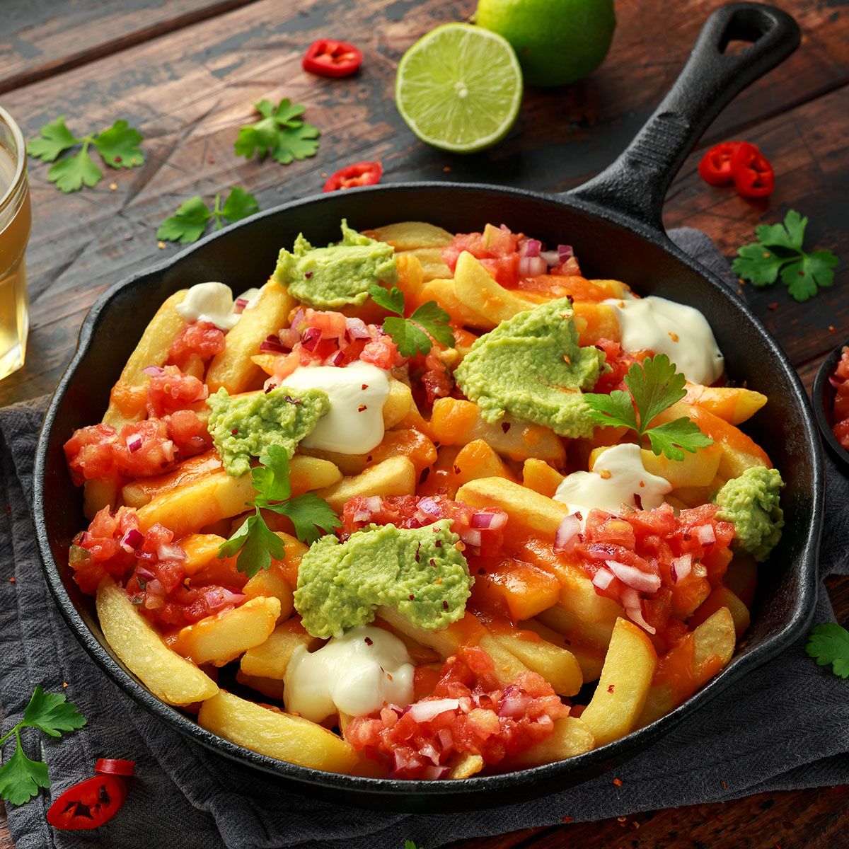 Pommes Frites mit Belag in einer schwarzen Pfanne. Enthält Guacamole, Tomatenwürfel und Soße. Limette und Bier daneben.