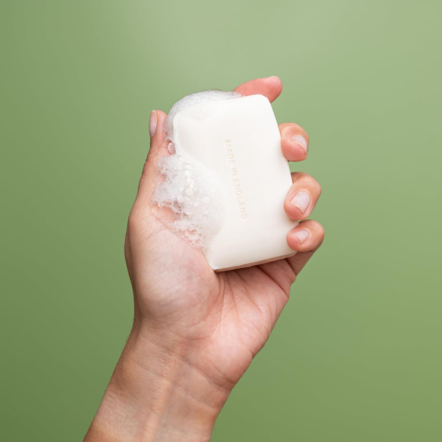 Hand hält Seife mit Schaum. Seife rechteckig, cremefarben. Text: Made in England.