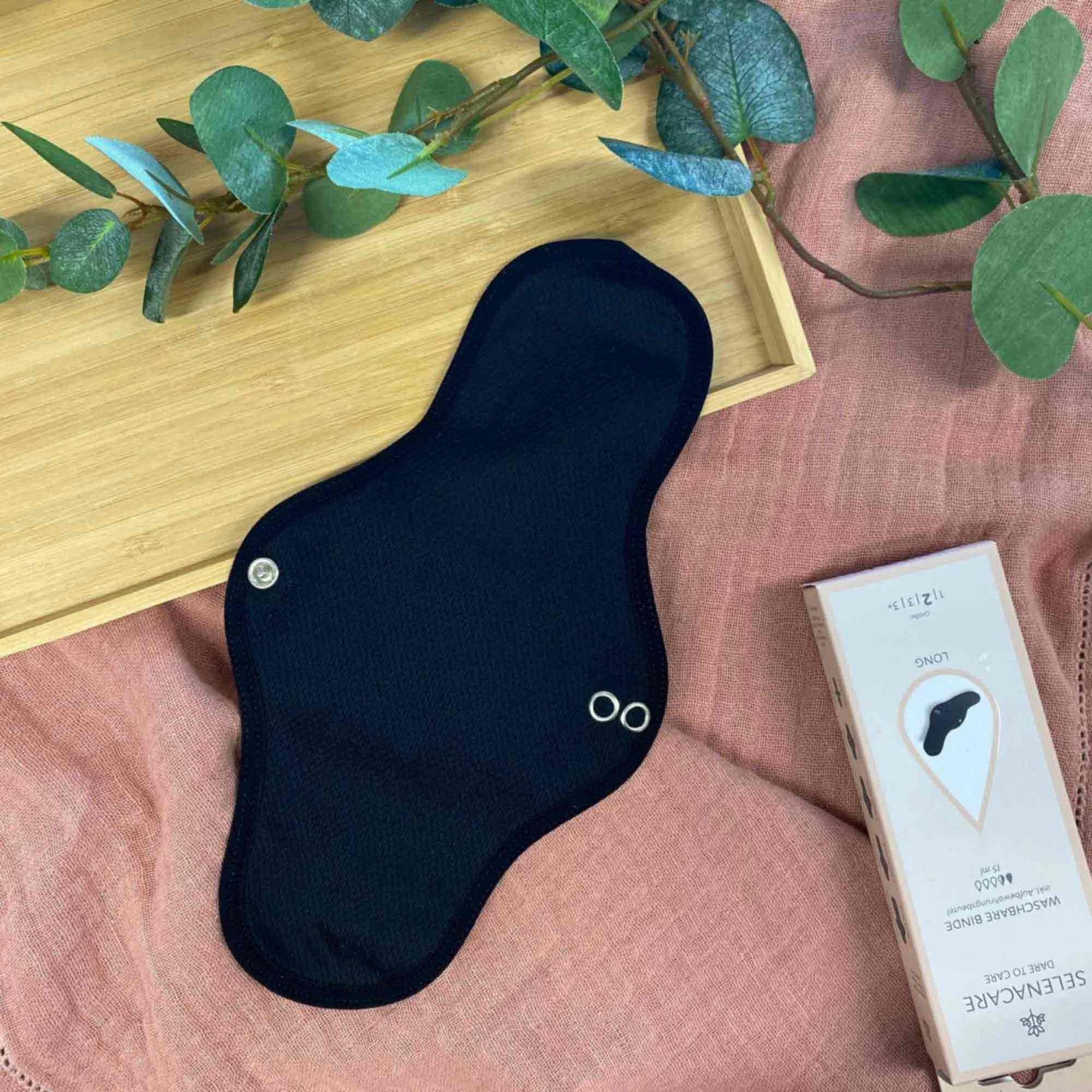 Schwarze, waschbare Binde auf rosa Stoff und Holztablett. Verpackung mit Logo und Produktinformationen. Binde mit Druckknöpfen und Logo.