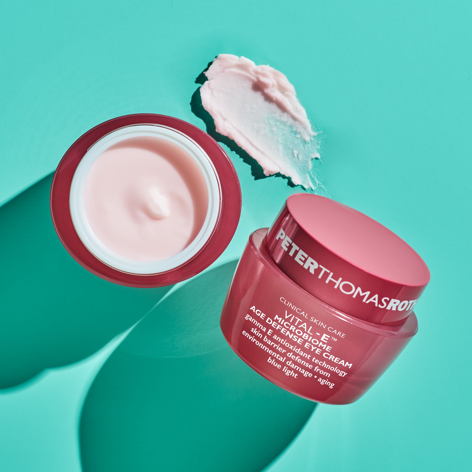 Offener rosa Tiegel mit Creme daneben. Deckel daneben. Text: VITAL-E MICROBIOME AGE DEFENSE EYE CREAM. Auf blauem Hintergrund.
