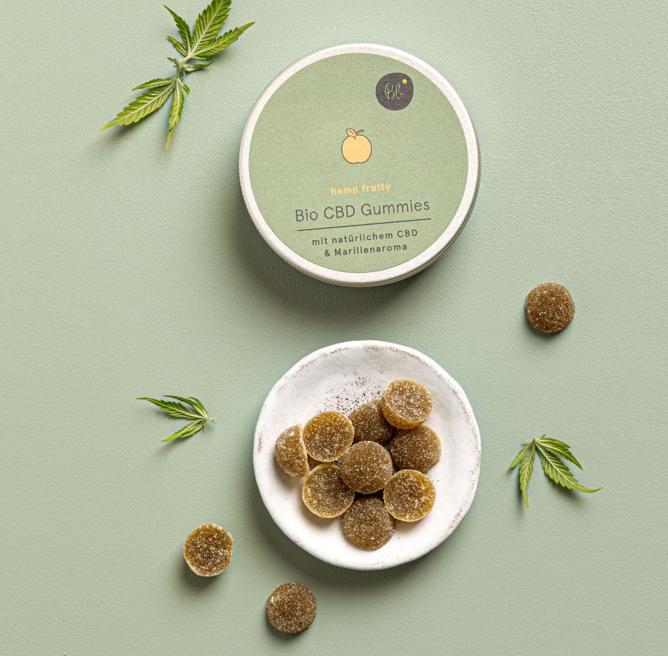 Geöffnete Dose "Bio CBD Gummies" mit Gummibärchen in Schale. Grüne Blätter liegen daneben.
