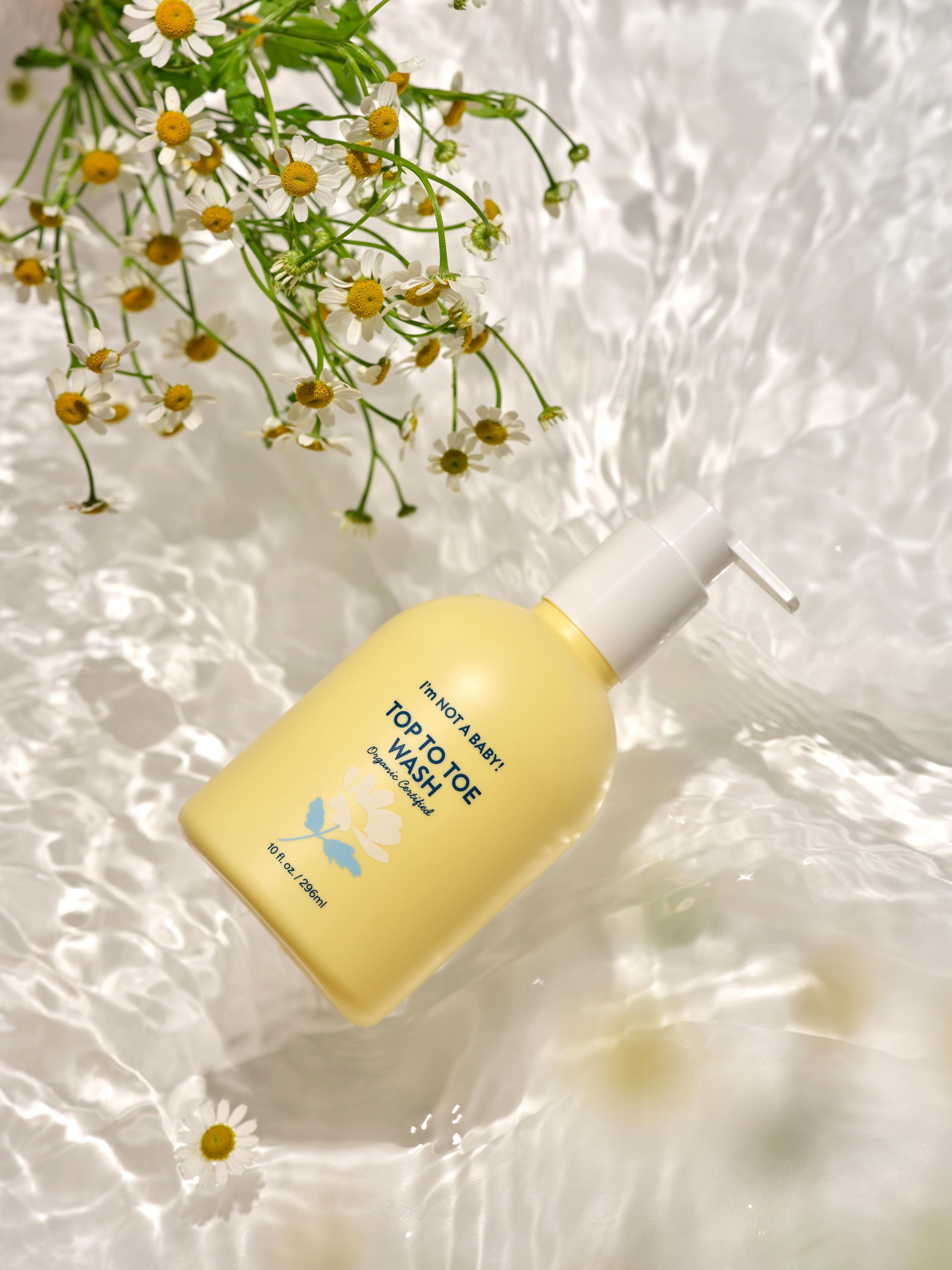 Gelbe Pumpflasche mit "TOP TO TOE WASH" Aufschrift, umgeben von Wasser und Kamillenblüten.