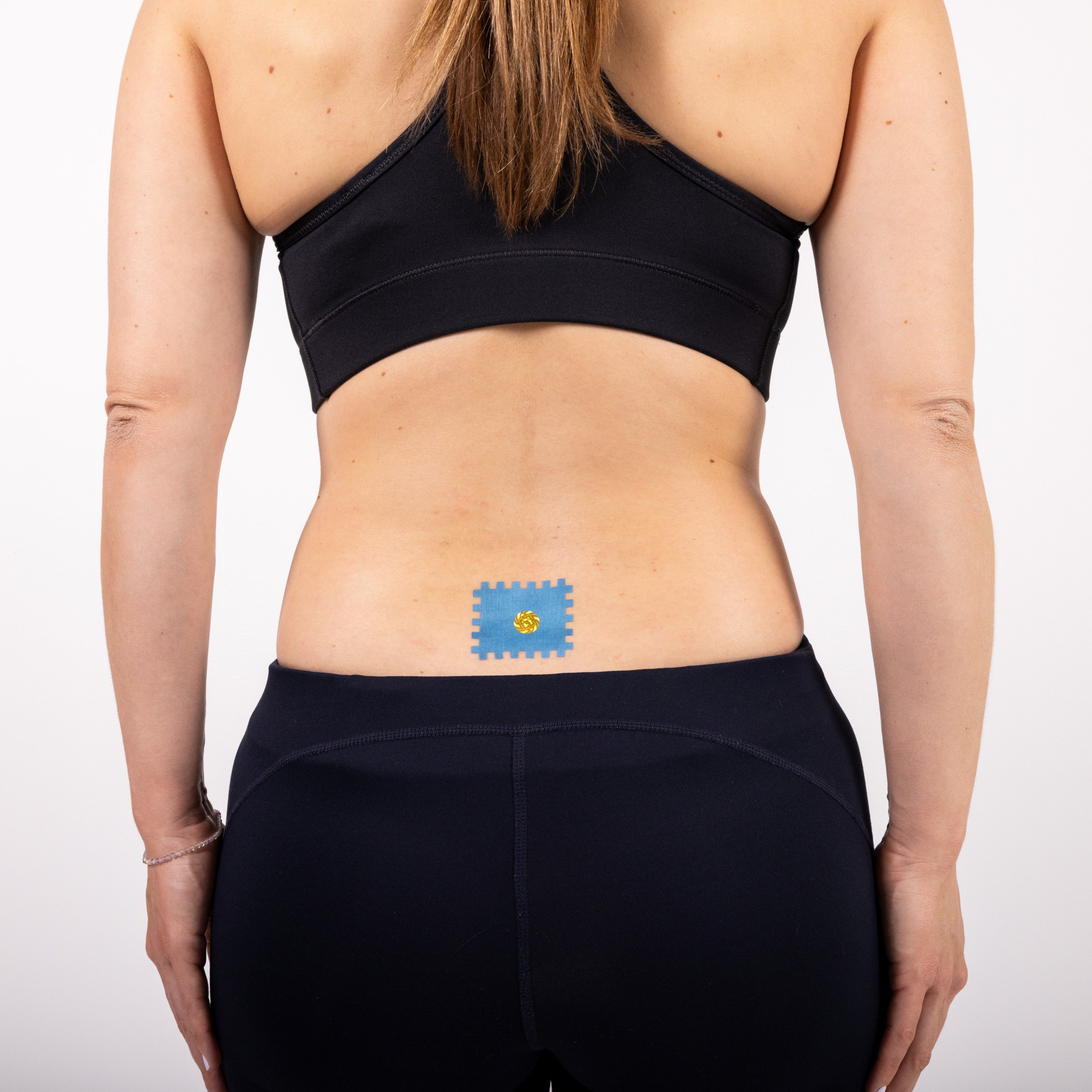 Rücken einer Frau mit einem blauen Pflaster auf dem unteren Rücken. Pflaster quadratisch mit gelbem Kreis. Frau trägt Sportkleidung.